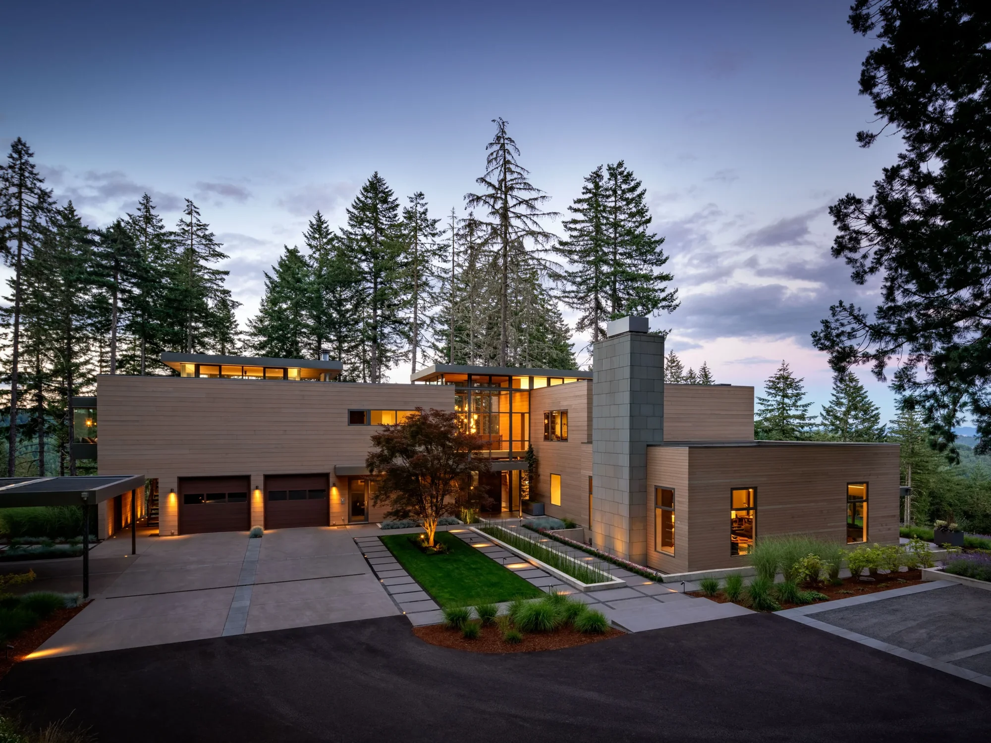 Modern two-story home with horizontal wood siding and stone tower, nestled among tall evergreen trees at dusk.