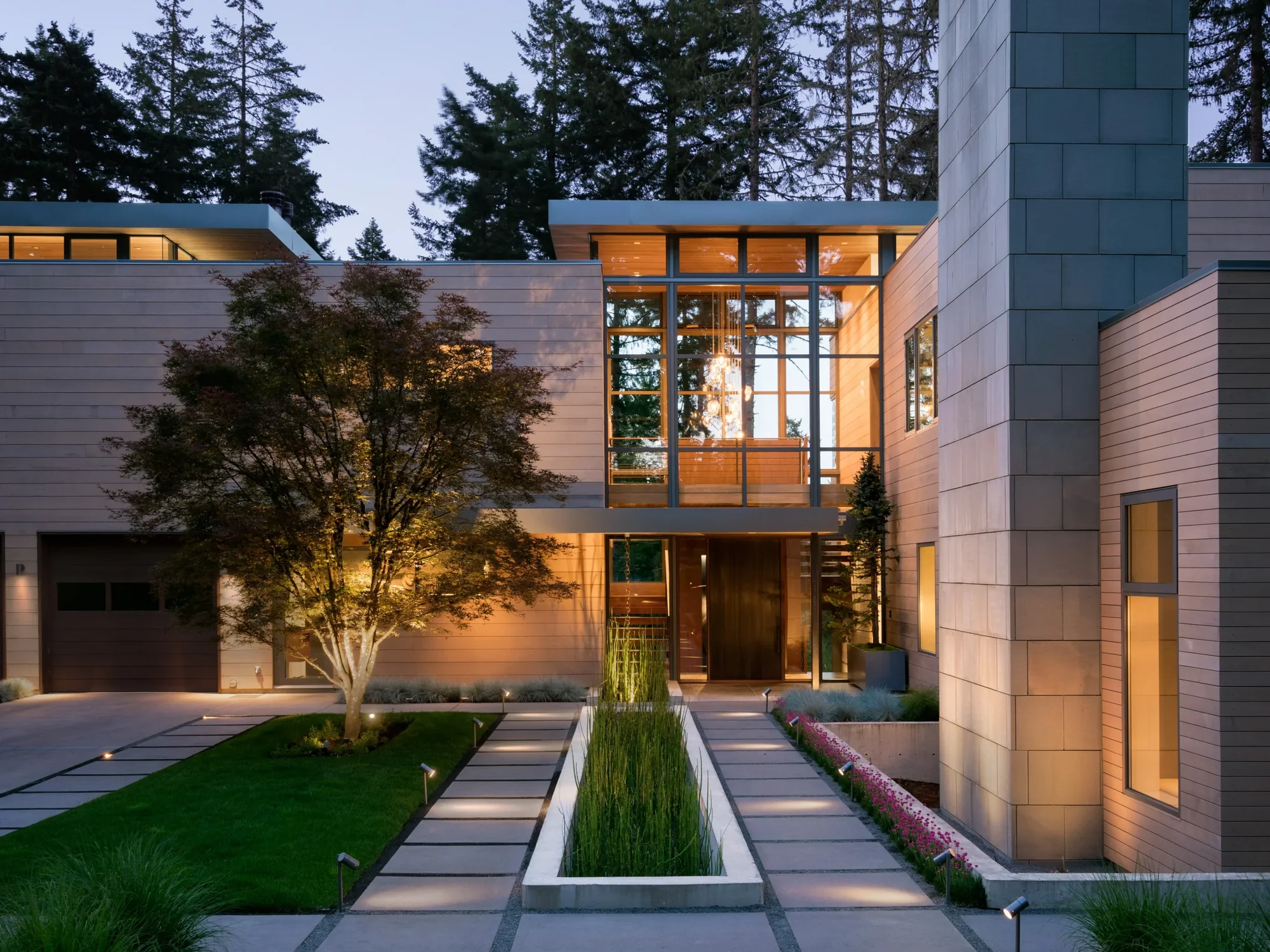 Modern two-story home with horizontal cedar siding and floor-to-ceiling windows at twilight, featuring illuminated interiors and landscaped entry.