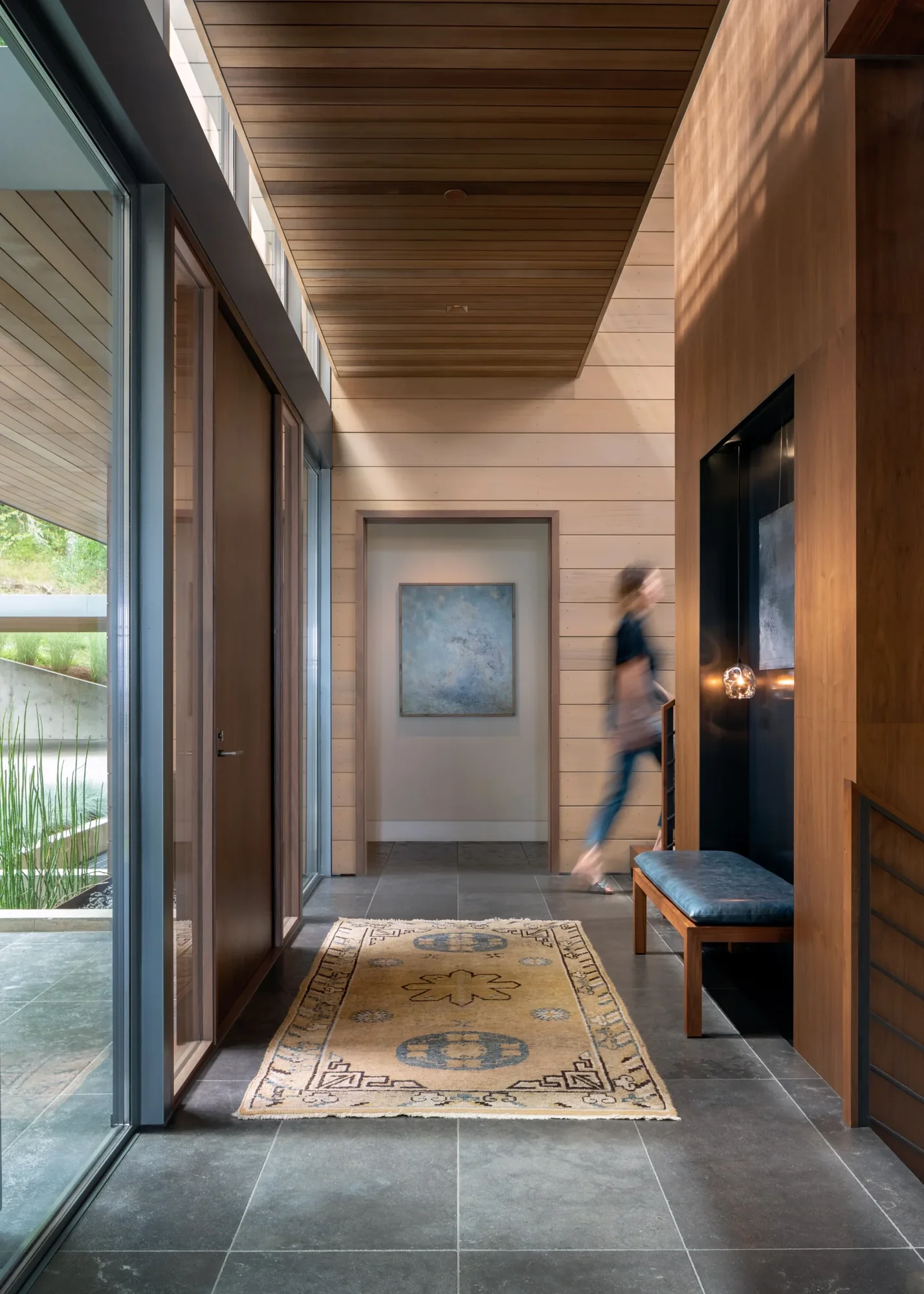 Modern hallway with wood ceiling, stone tile floor, vintage rug, and blurred figure walking past framed artwork