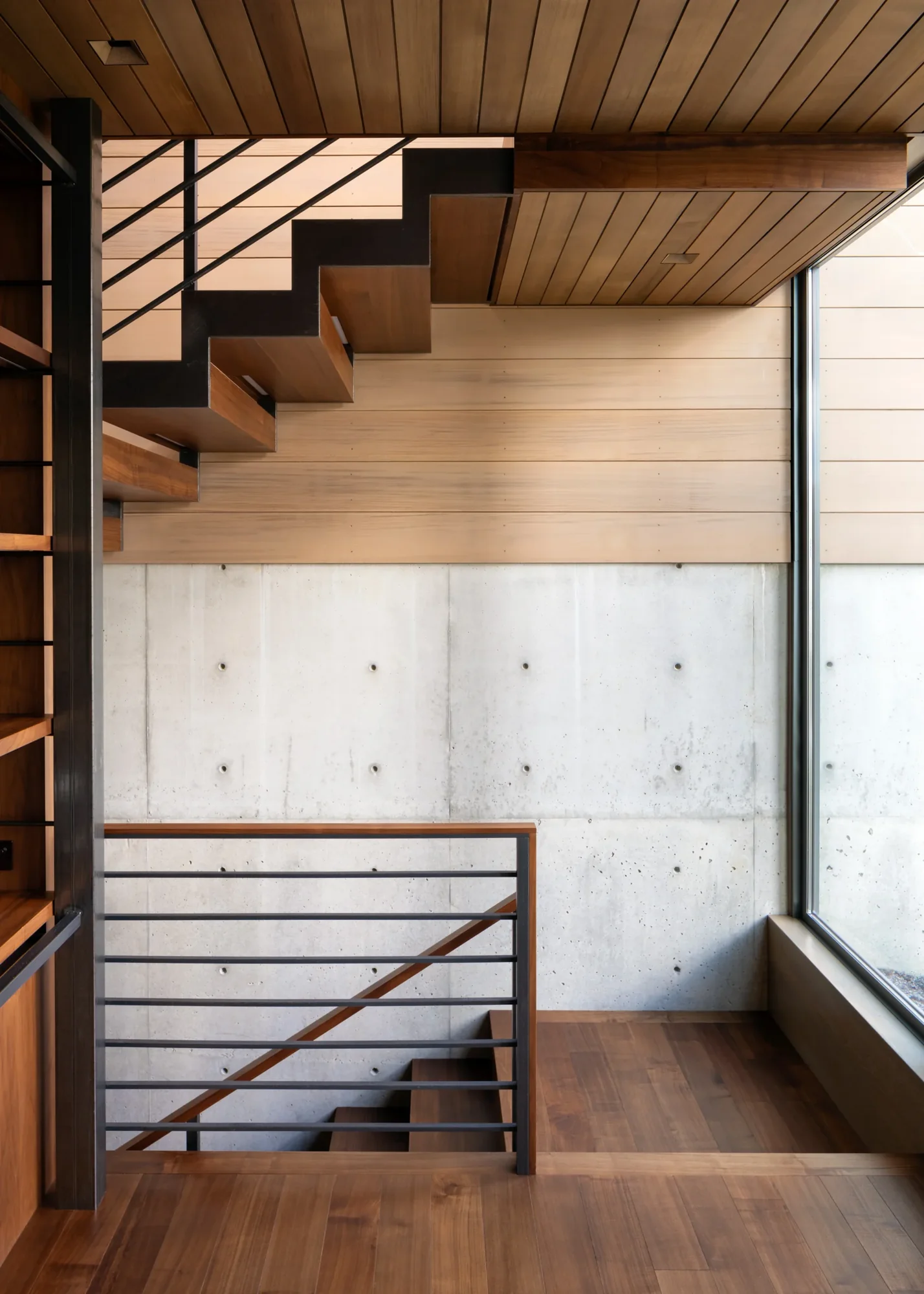 Modern staircase with wood treads and metal railings against concrete wall, featuring wood-paneled ceiling and natural light.