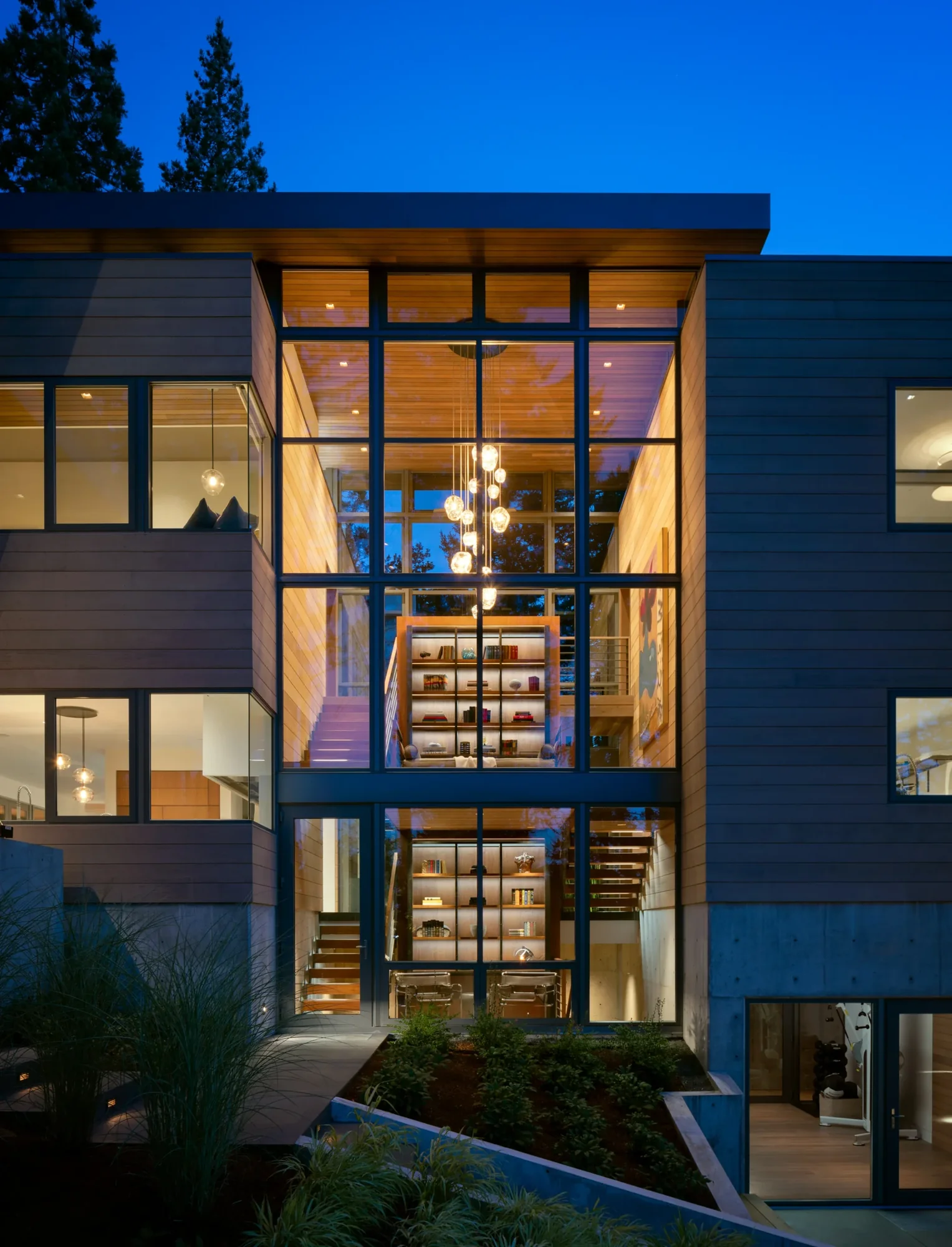 Modern multi-story home at dusk with floor-to-ceiling windows revealing illuminated interiors and built-in shelving throughout.