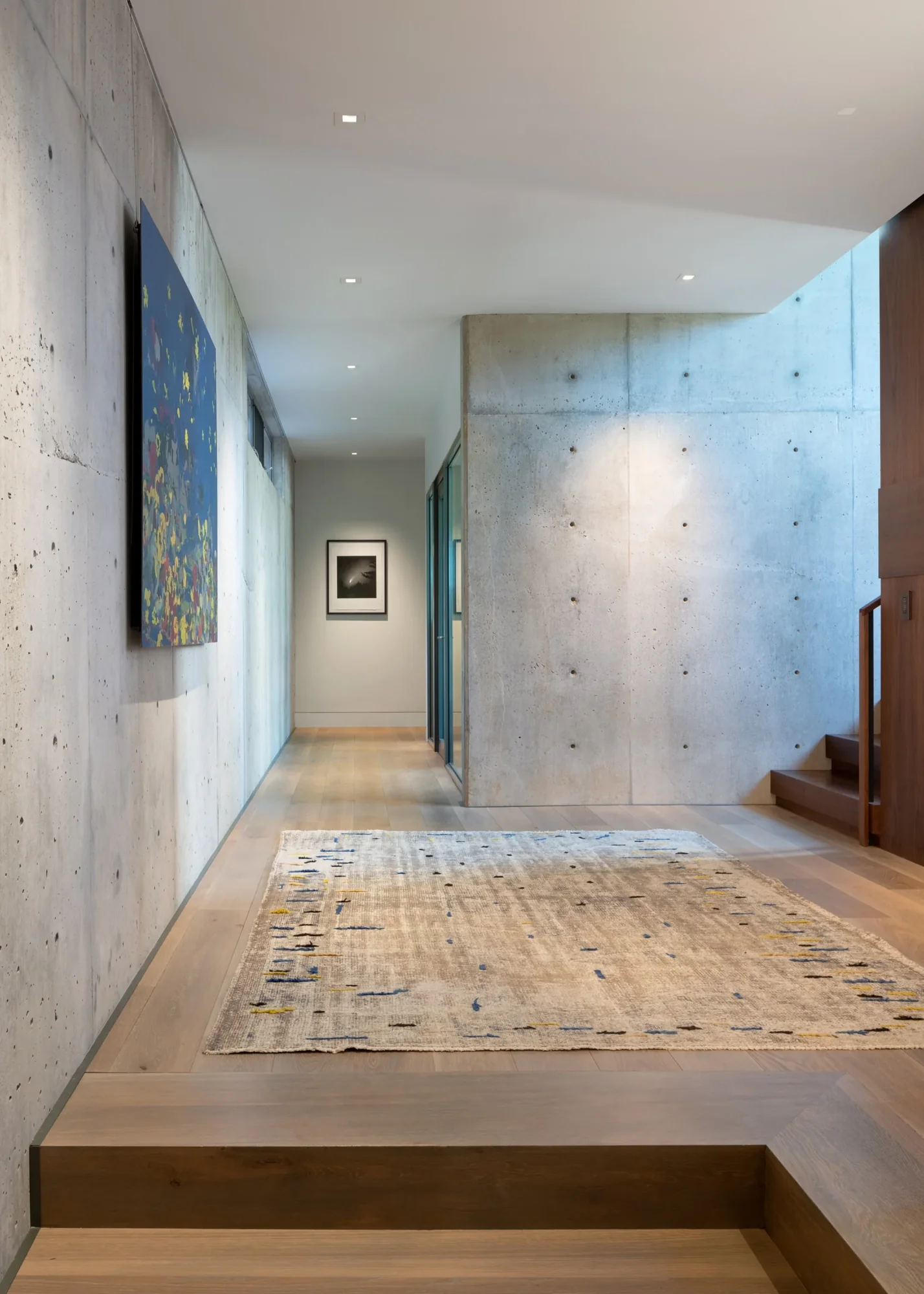 Modern hallway with concrete walls, light wood flooring, contemporary art, and woven area rug connecting interior spaces