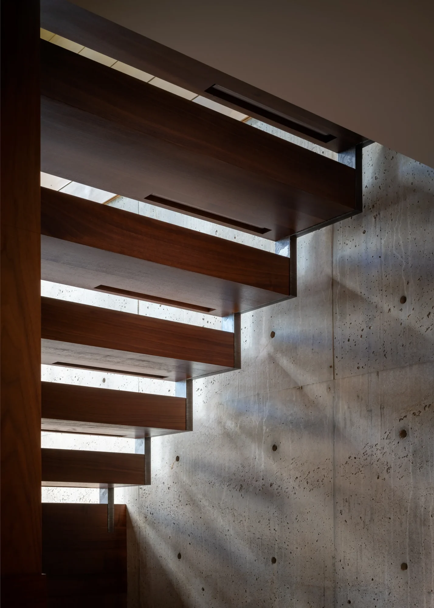 Floating wooden staircase against textured concrete wall with natural light filtering through horizontal gaps
