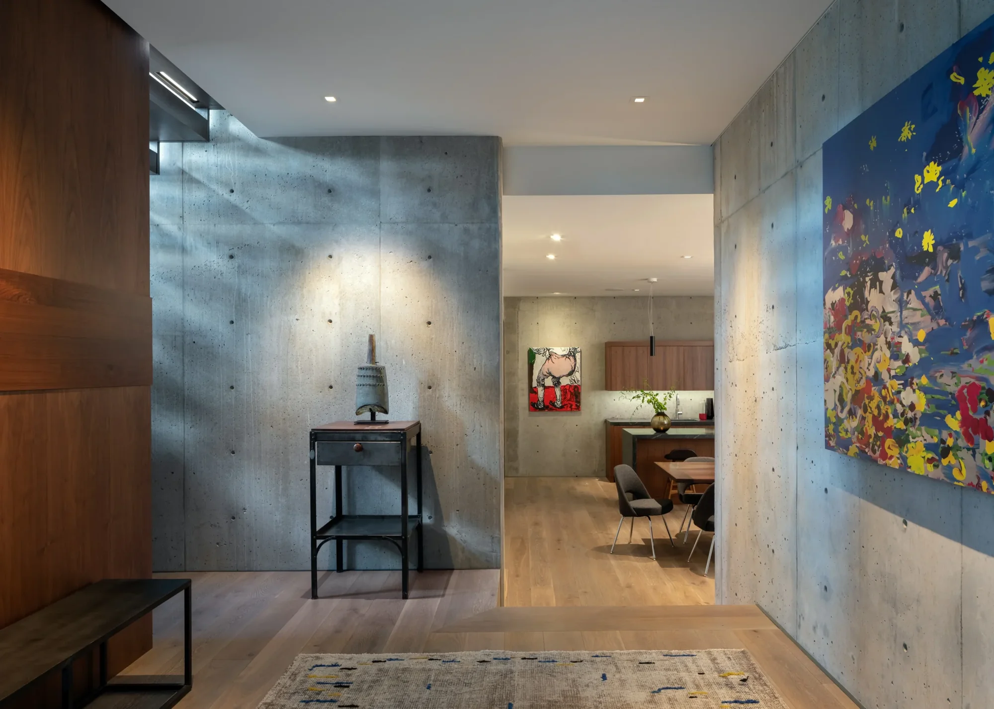 Modern interior hallway with exposed concrete walls, light wood flooring, and colorful artwork leading to kitchen area