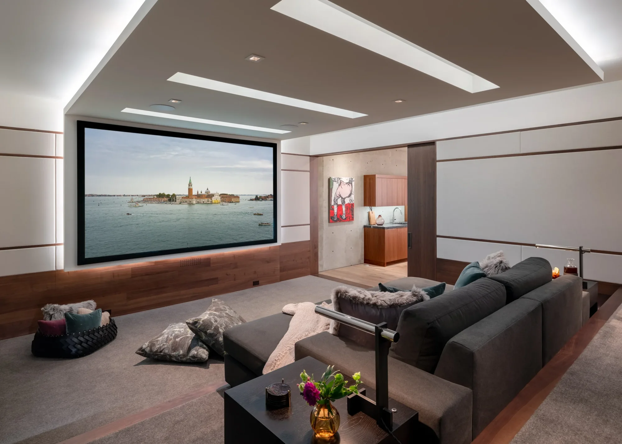 Modern home theater with large projection screen, dark gray sectional sofa, and illuminated wood platform base.