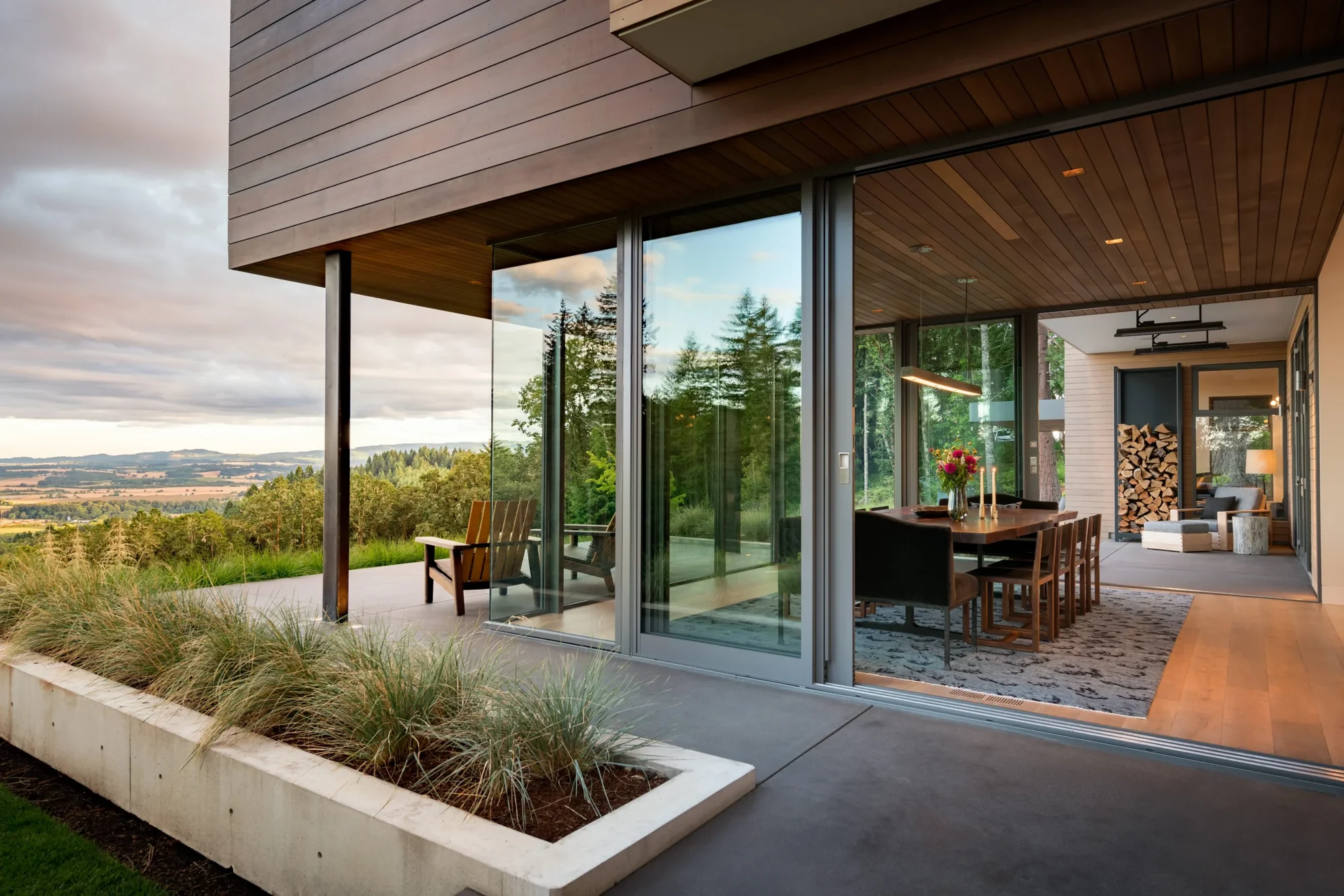 Modern home with floor-to-ceiling glass doors opening dining area to covered patio, overlooking scenic valley landscape.