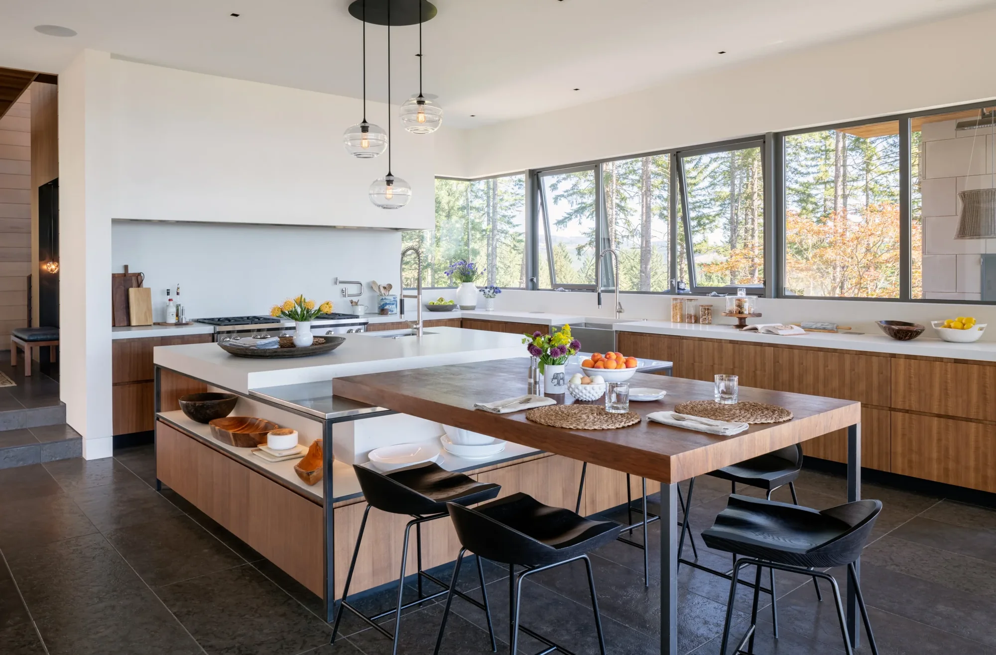 Modern kitchen with walnut cabinetry, white countertops, and large windows overlooking trees, featuring integrated island seating.