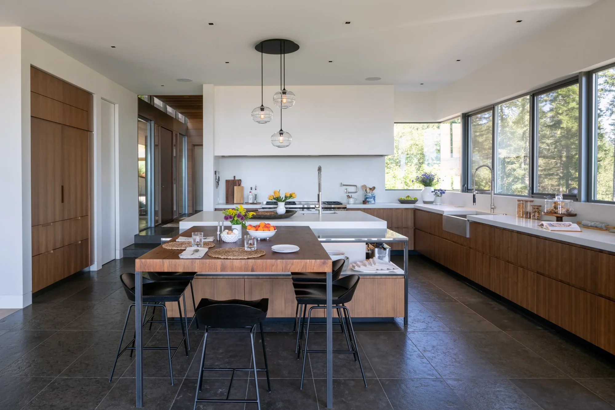Modern kitchen with wood cabinetry, white countertops, pendant lighting, and floor-to-ceiling windows overlooking trees.