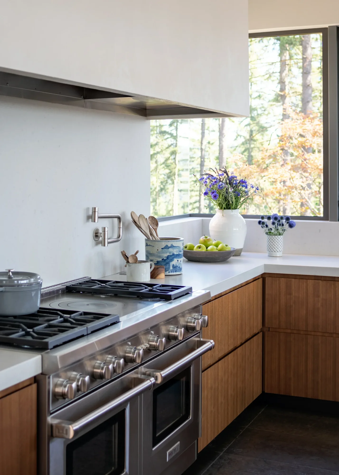 Modern kitchen with gas range, white countertops, wood cabinets, and large window framing forest views