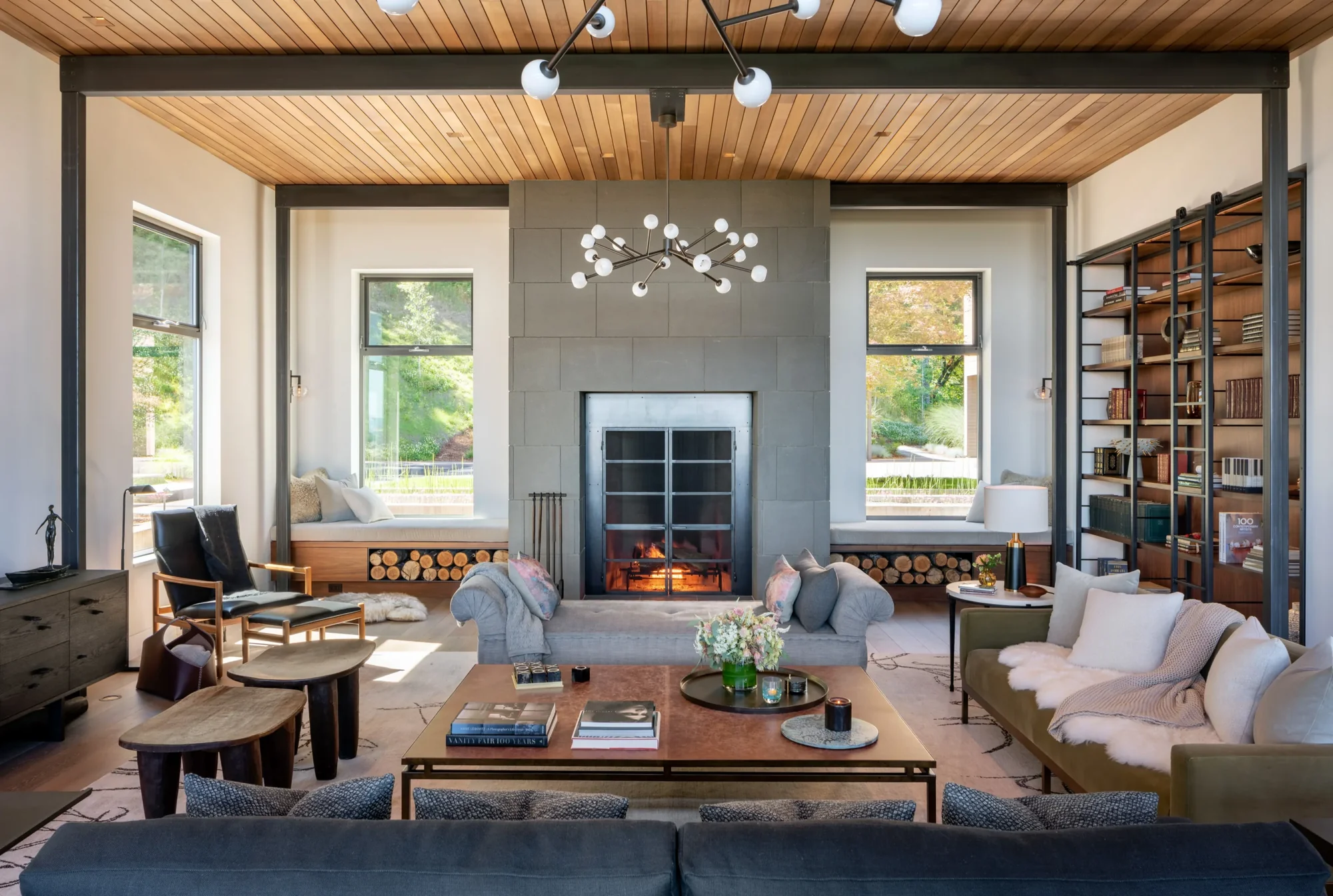 Modern living room with gray stone fireplace, wood slat ceiling, window seat nooks, and indoor-outdoor connection.