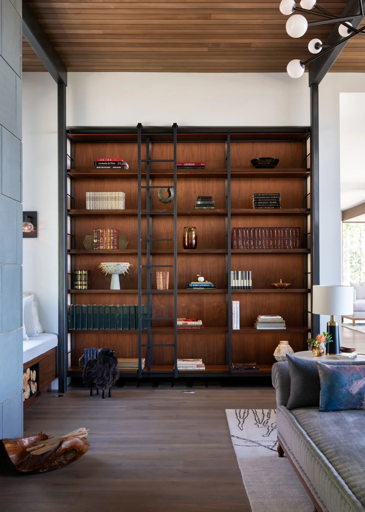 Floor-to-ceiling walnut bookshelf with black metal ladder in living room beneath wood-paneled ceiling