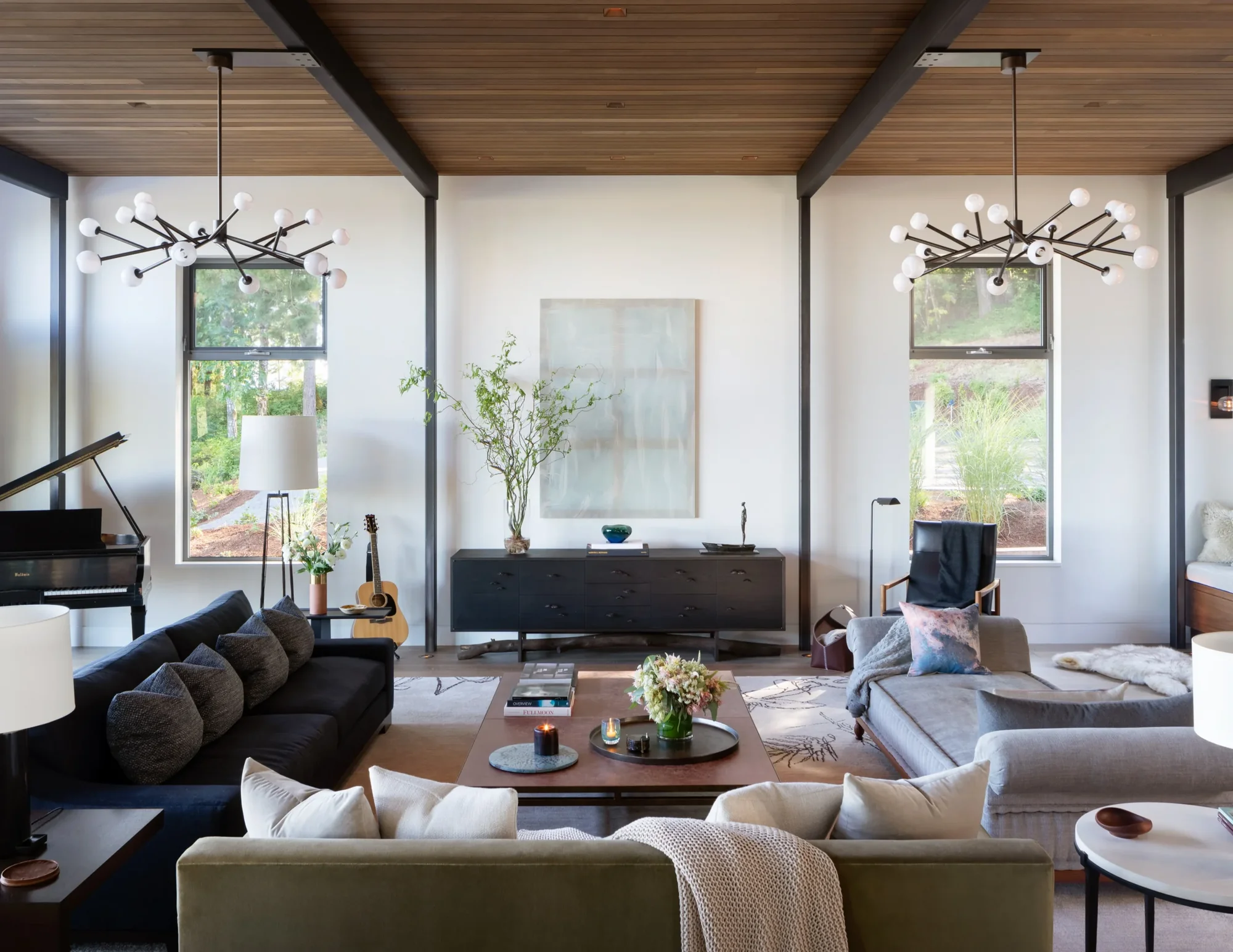 Modern living room with wood ceiling, black steel beams, sputnik chandeliers, and windows overlooking natural landscape