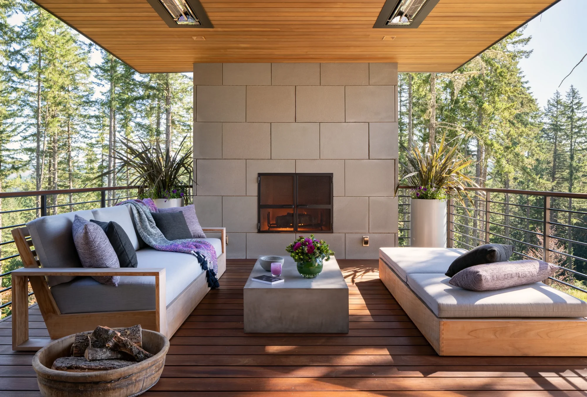 Covered outdoor deck with fireplace, modern furniture, and forest views framed by wood ceiling and cable railing