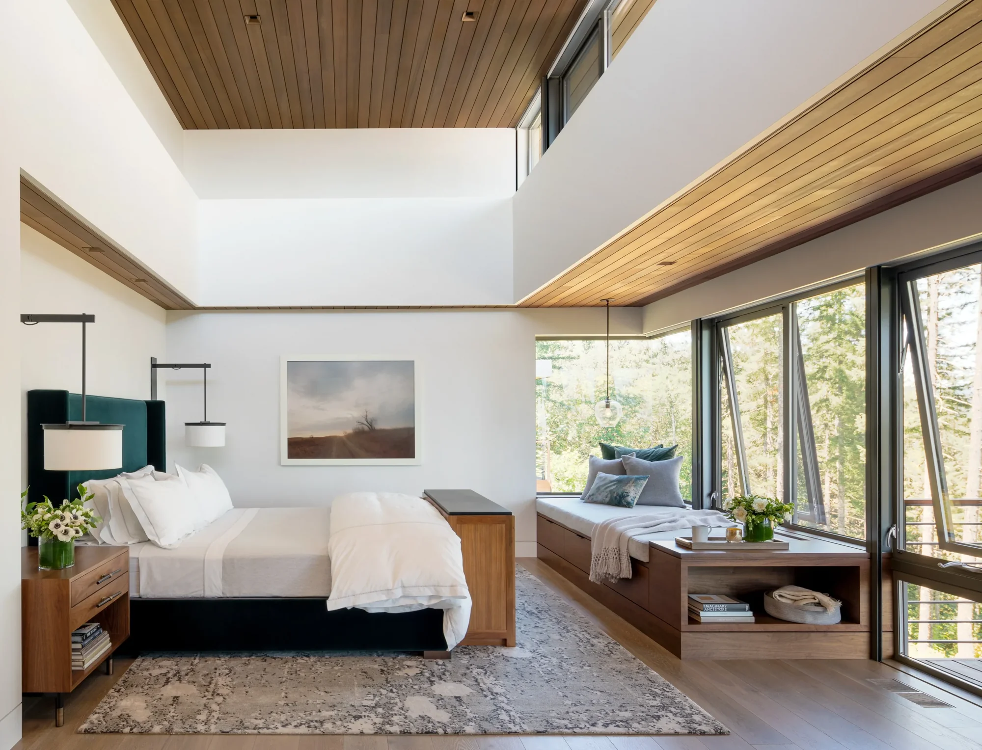 Modern bedroom with wood ceiling, floor-to-ceiling windows, built-in window seat, and views of surrounding trees.