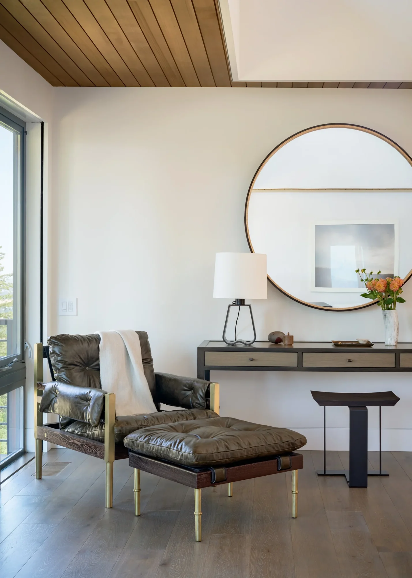 Modern reading nook with leather chair and ottoman, wood ceiling, circular mirror, and console table near window