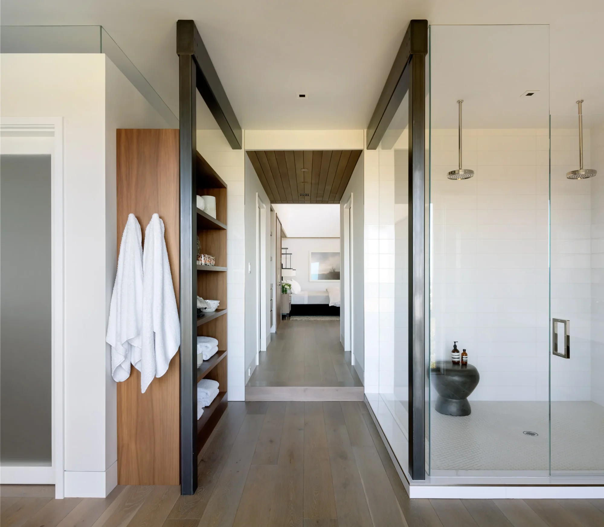 Modern bathroom hallway with wood shelving, glass shower enclosure, and view through to bedroom beyond