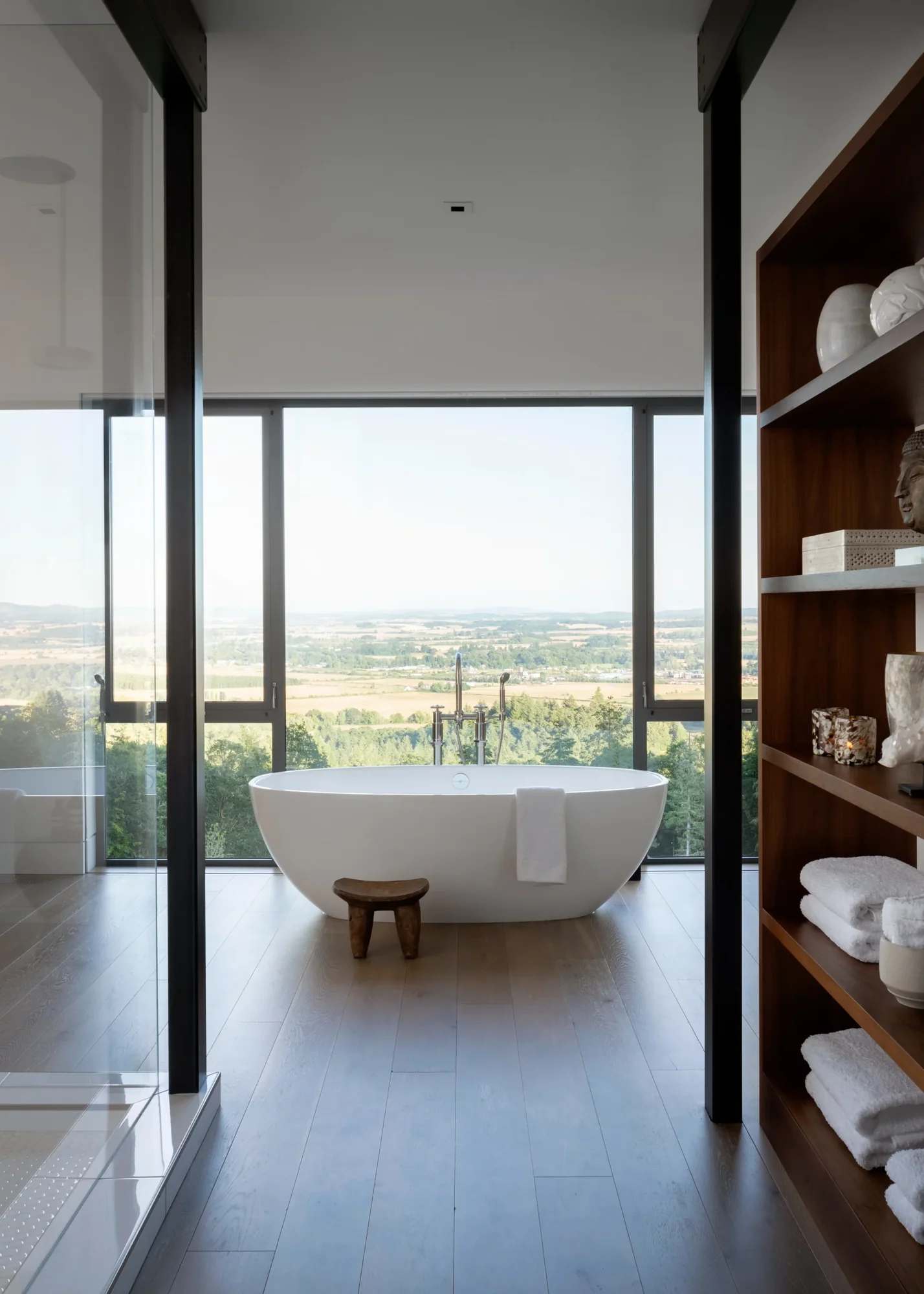 Modern bathroom with freestanding white tub, floor-to-ceiling windows overlooking valley, and walnut shelving with towels.