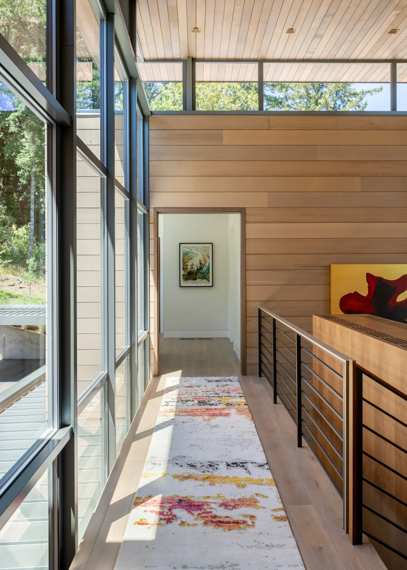 Modern hallway with horizontal cedar siding, clerestory windows, colorful area rug, and views to outdoor deck and greenery