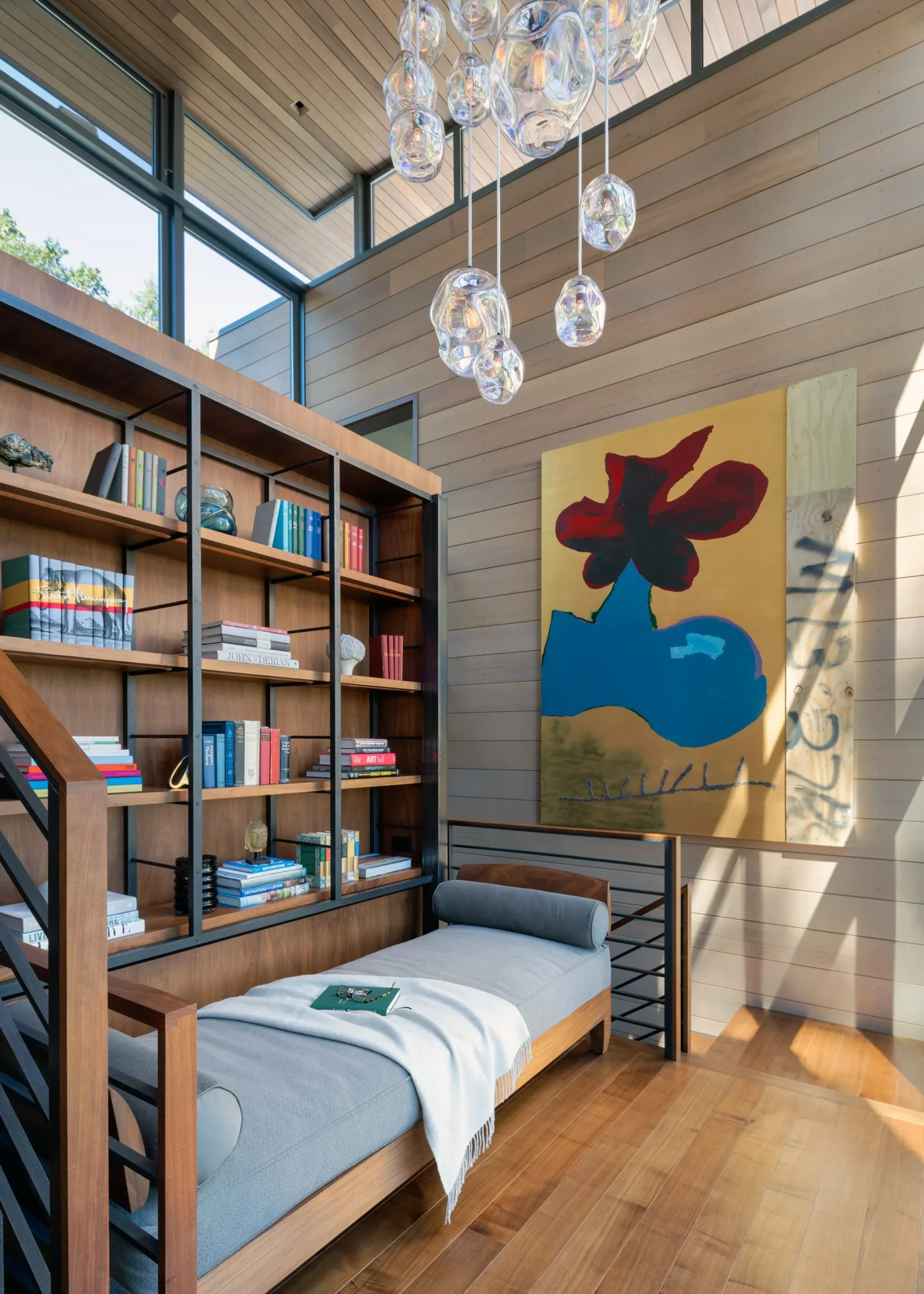 Modern reading nook with wood built-in shelving, daybed, abstract artwork, and glass pendant lighting in high-ceilinged space