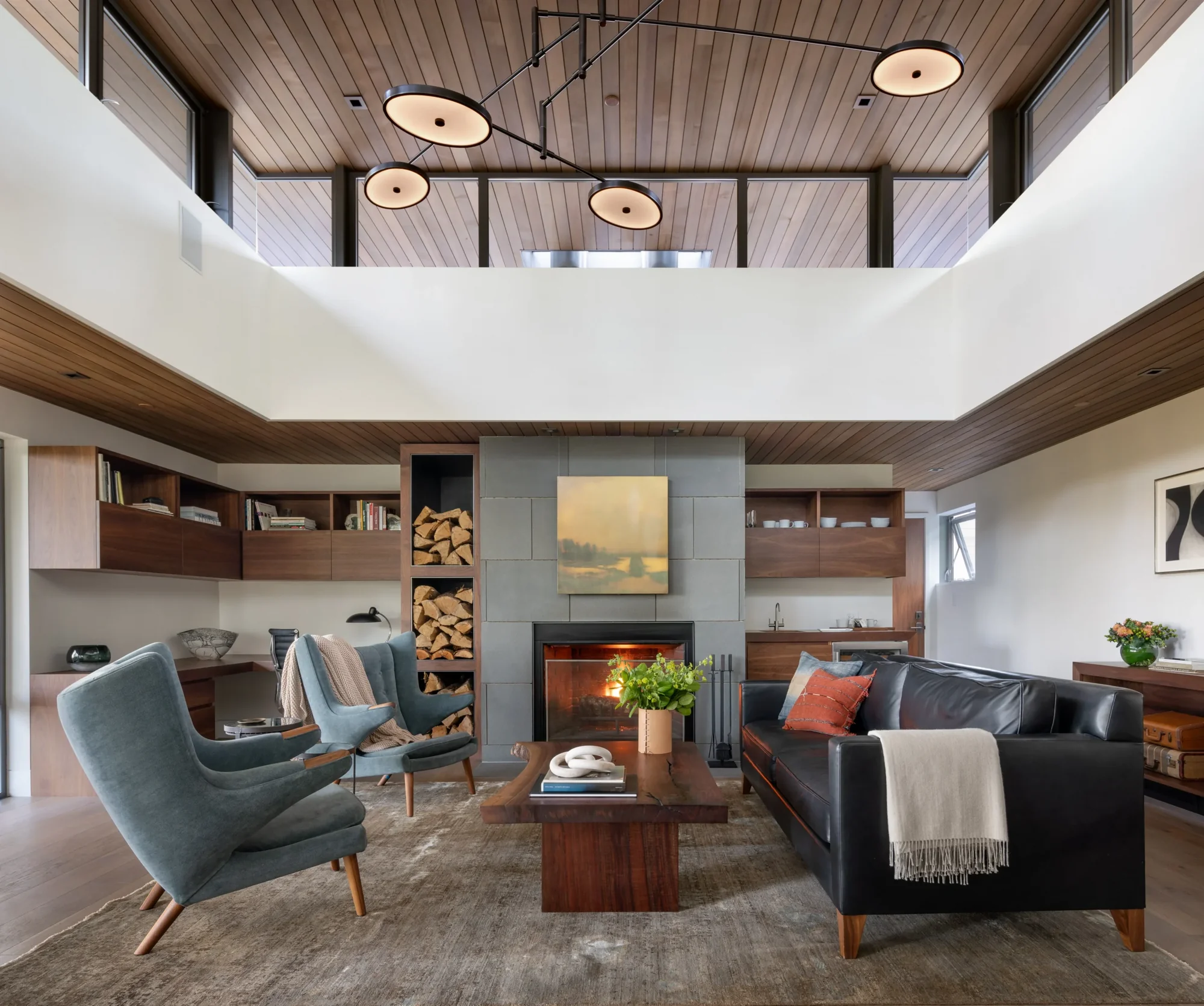Modern living room with wood ceiling, fireplace, blue armchairs, black leather sofa, and double-height space with upper-level views.