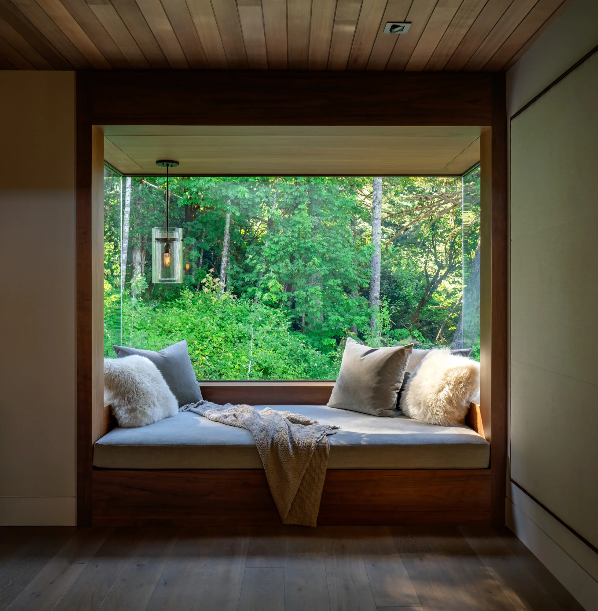 Cozy window seat nook with cushions and fur pillows overlooking lush green forest through large picture window.