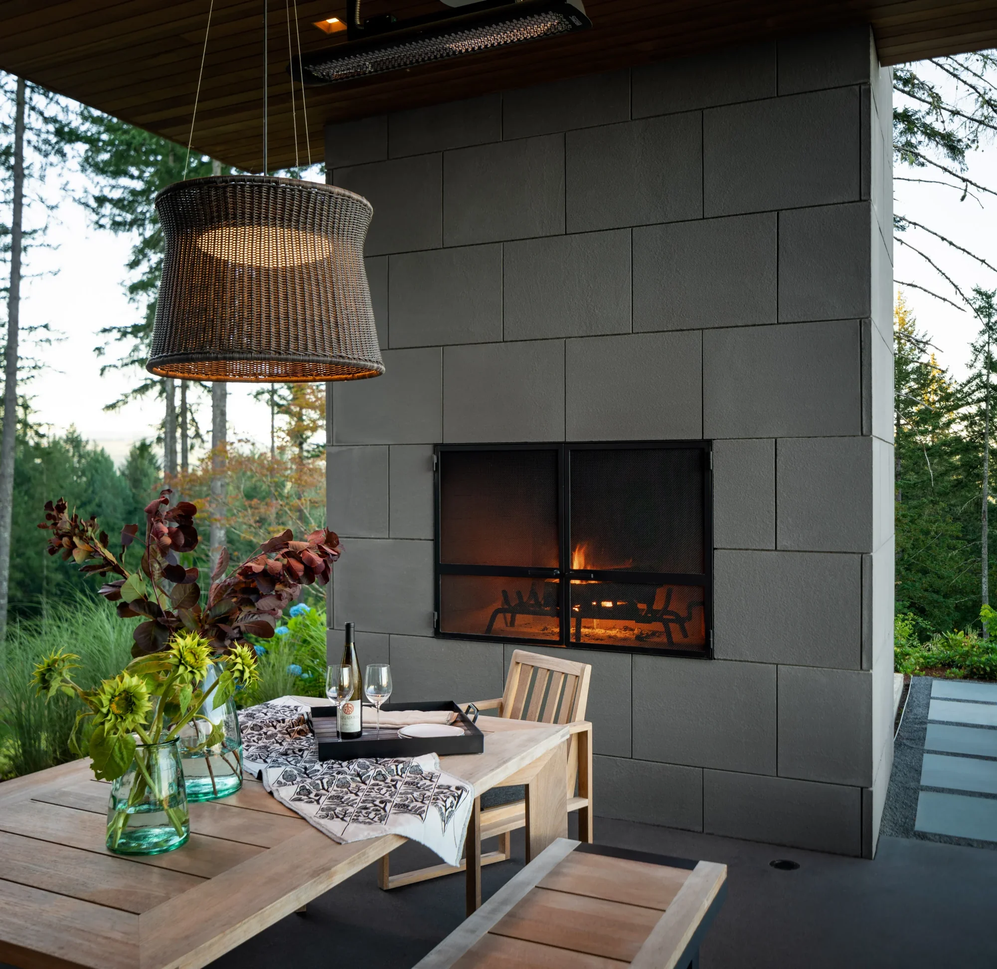 Covered outdoor dining area with fireplace, wicker pendant light, and natural wood table overlooking forest.