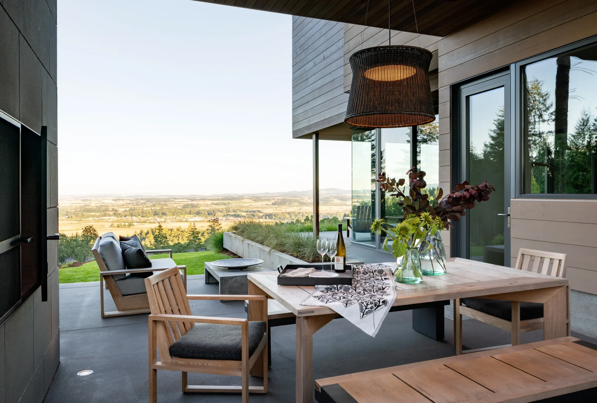 Modern covered patio with wooden dining furniture, woven pendant light, and panoramic valley views framed by glass doors