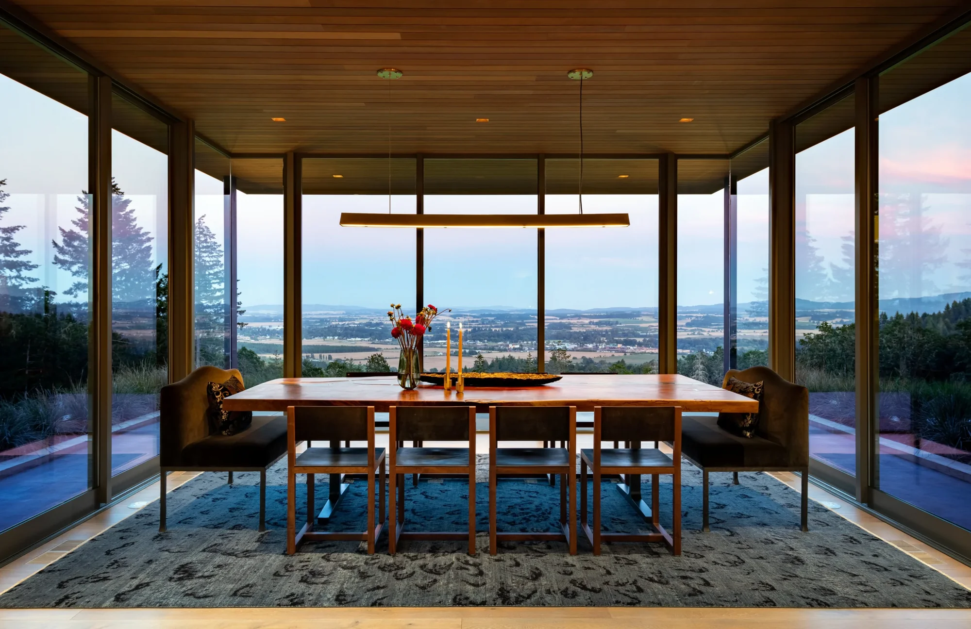 Modern dining room with floor-to-ceiling windows, wood table and chairs, panoramic valley views, and warm wood ceiling.