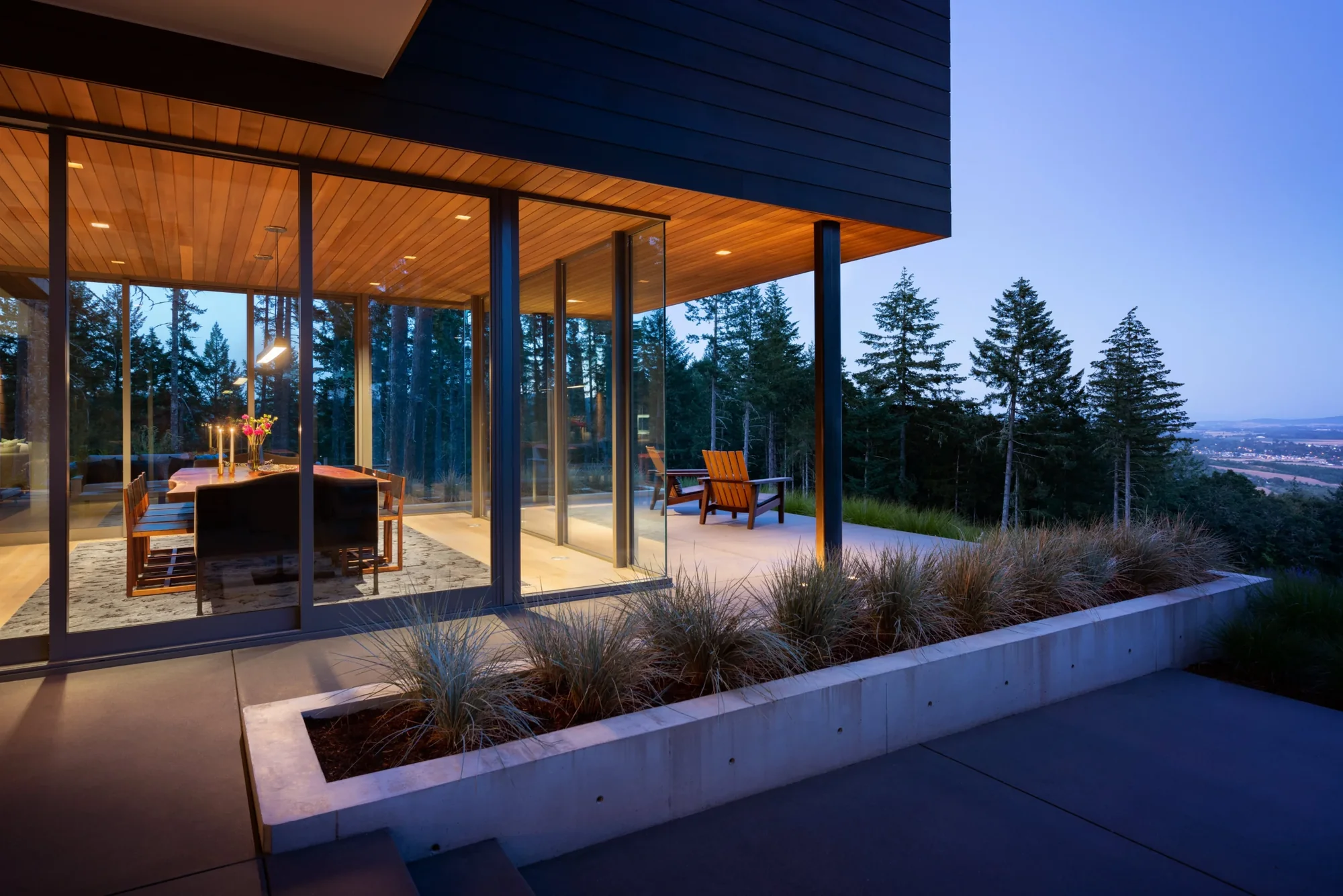 Modern home with floor-to-ceiling glass walls opening to hillside patio, featuring cedar ceiling and mountain views at dusk.