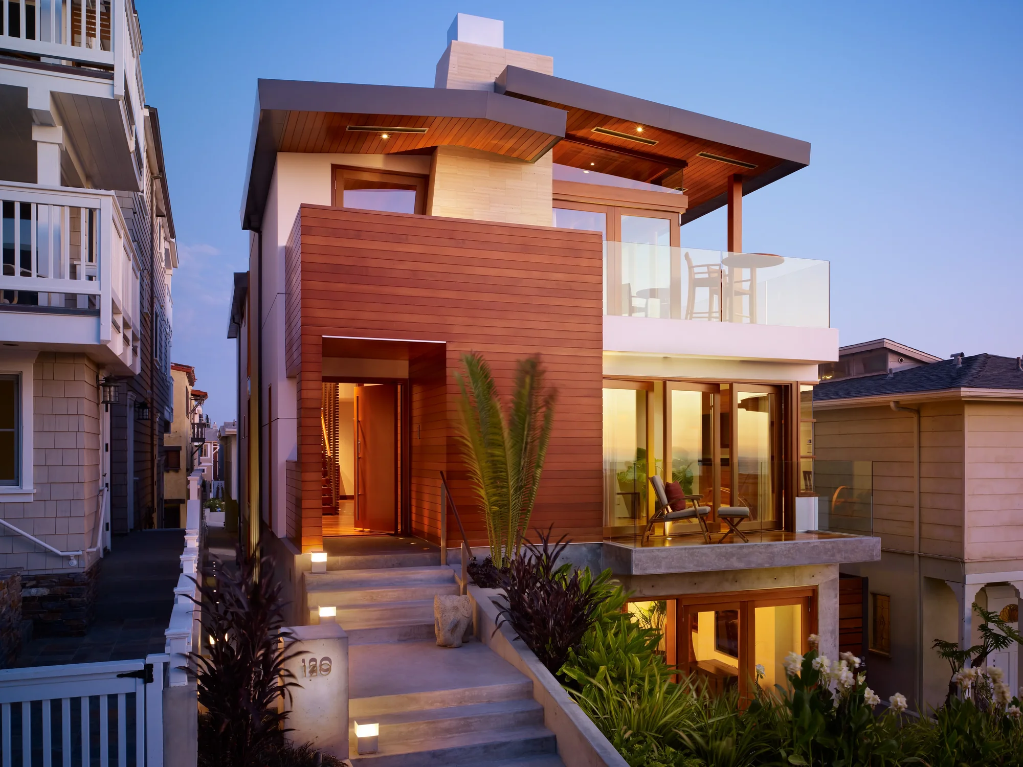 Modern three-story beach house with cedar siding, rooftop deck, and illuminated interiors at dusk