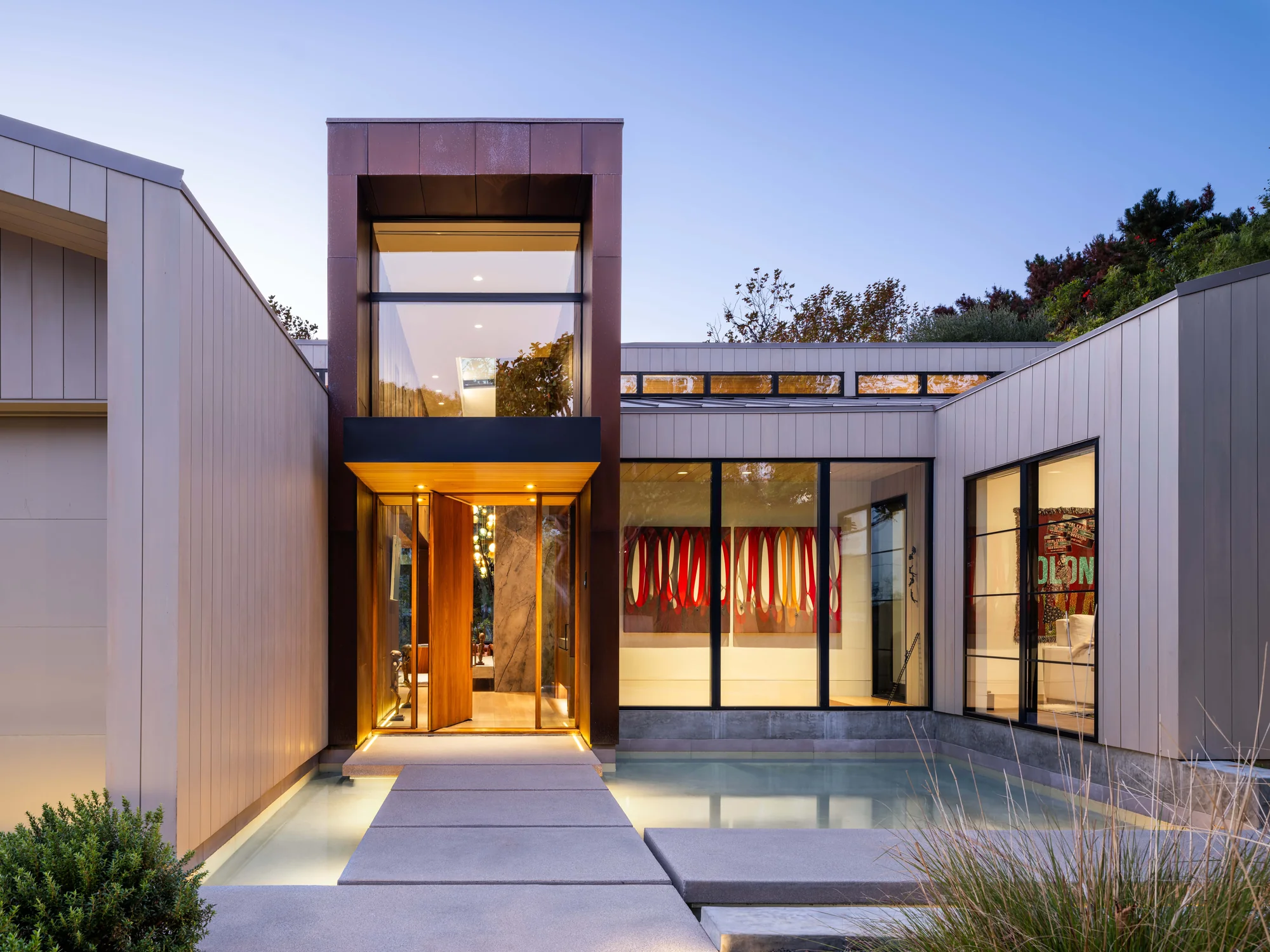 Modern home entrance with floor-to-ceiling glass, weathered steel tower, and illuminated pathway at dusk