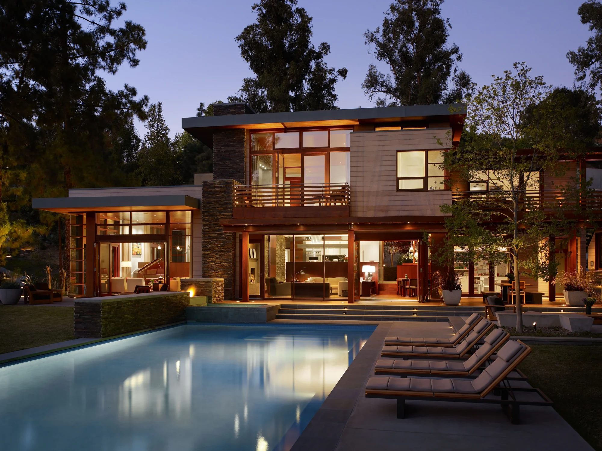 Twilight view of a custom modern home with mahogany framing, stone cladding, open living spaces, and a illuminated pool.