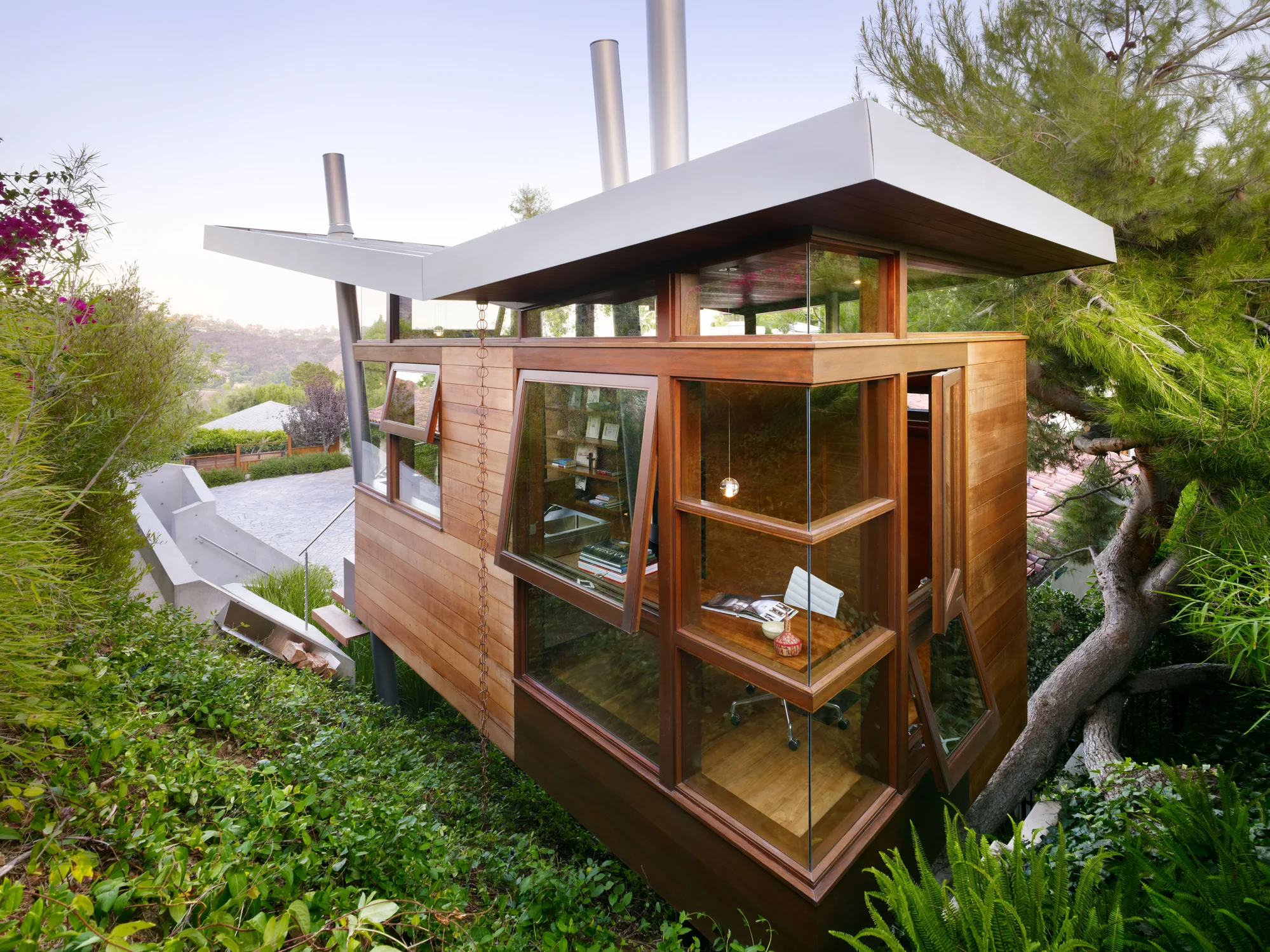 Modern cedar-clad home office with floor-to-ceiling glass walls, white cantilevered roof, and lush hillside garden integration.