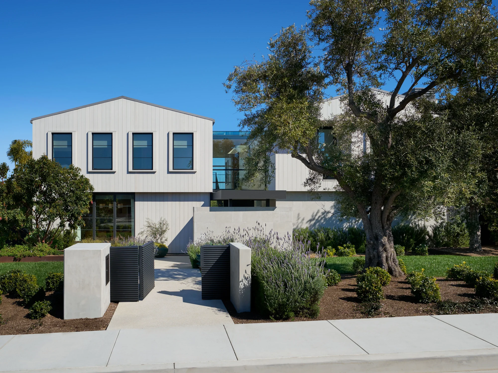 Modern white two-story home with black-framed windows, surrounded by mature trees and drought-tolerant landscaping.