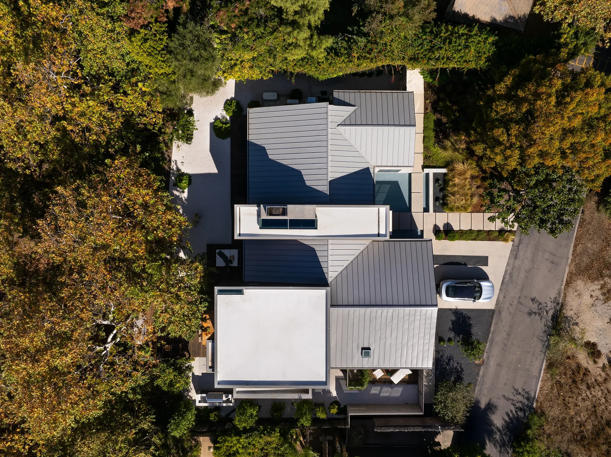 Aerial view of modern home with gray roof and white walls nestled among trees, featuring outdoor terraces and driveway.