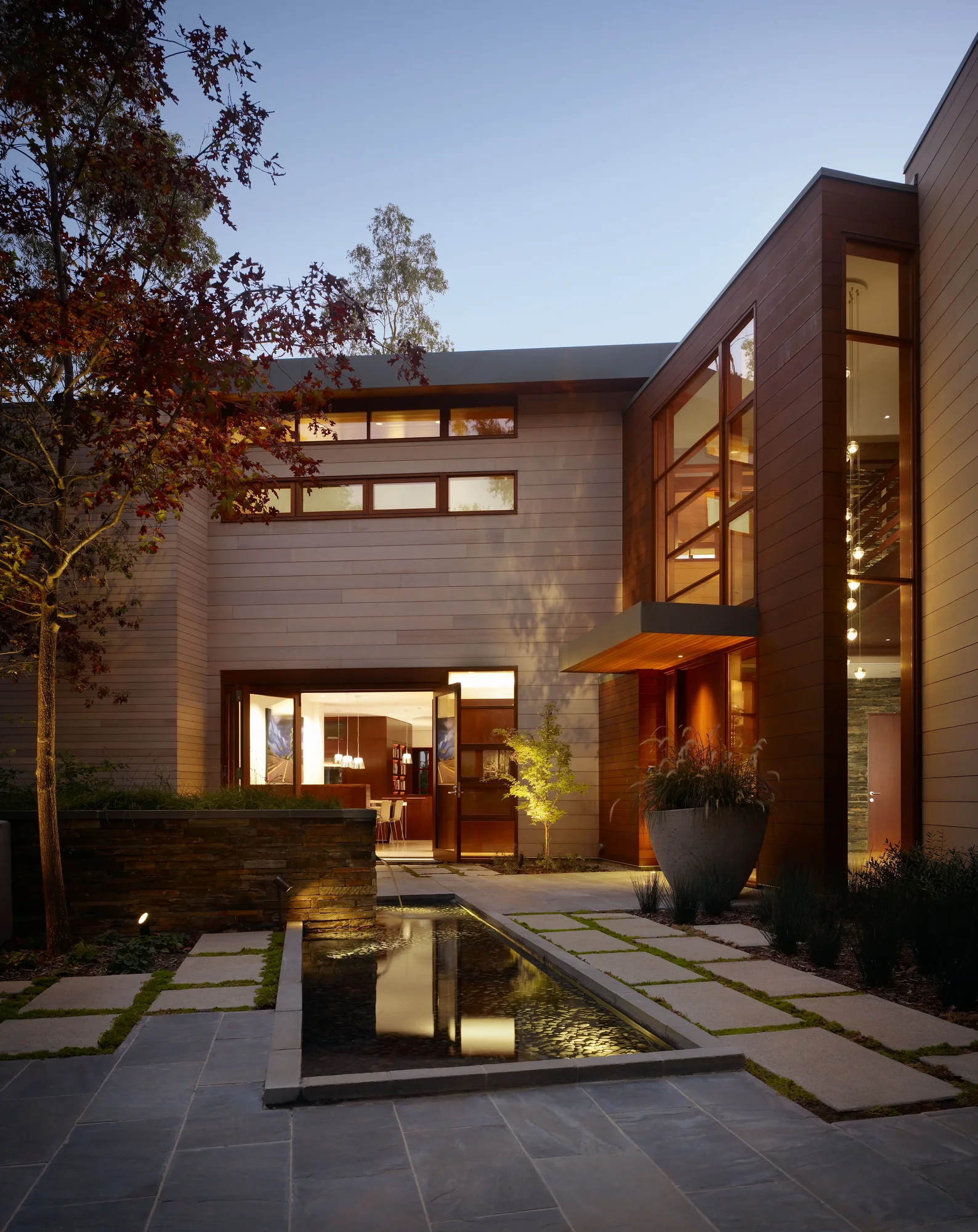 Twilight exterior of a custom home with warm mahogany cladding, floor-to-ceiling glazing, and a reflective entry water feature.