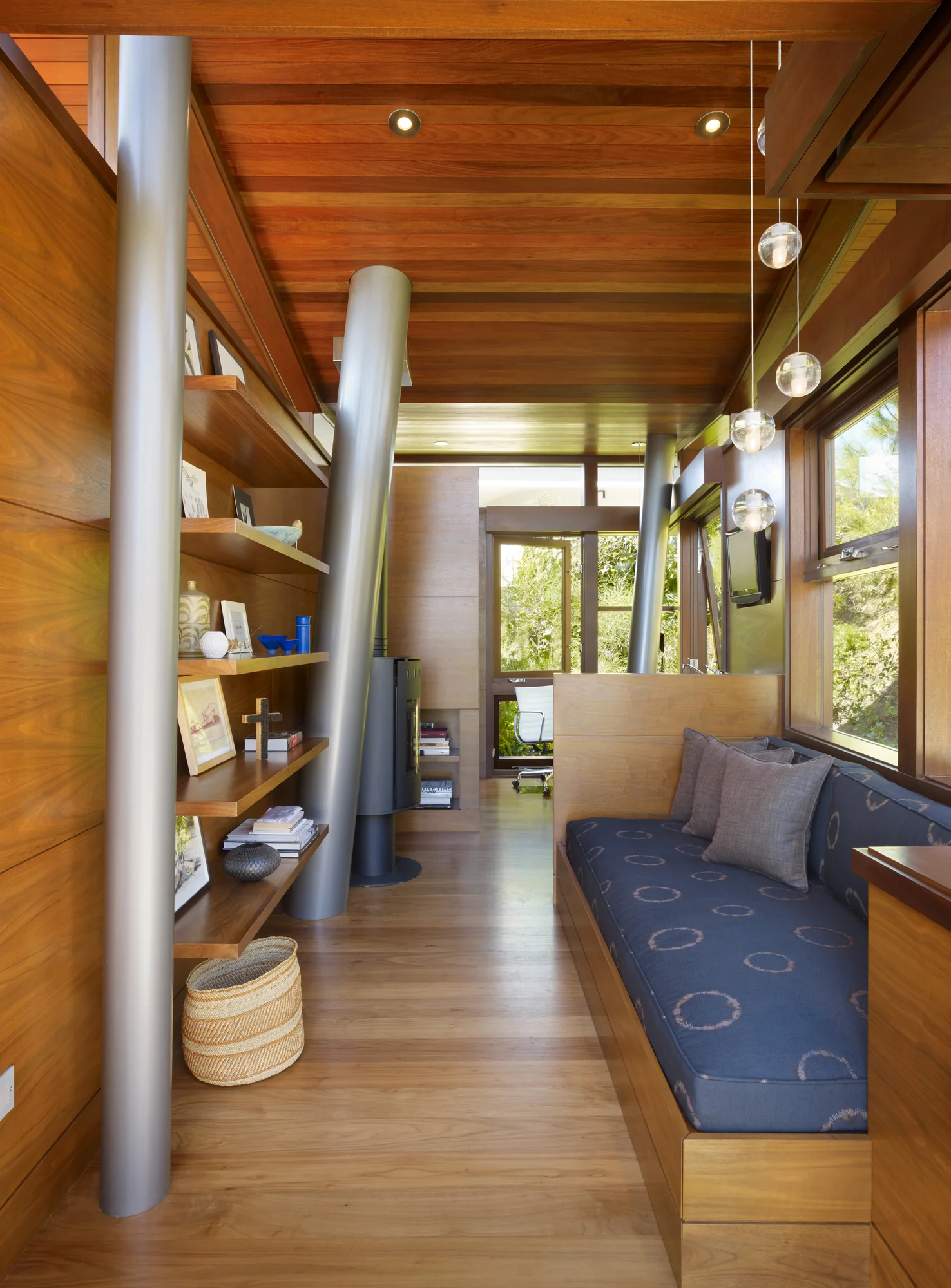 Wood-paneled reading nook with built-in bench seating, open shelving, cylindrical steel columns, and pendant lighting