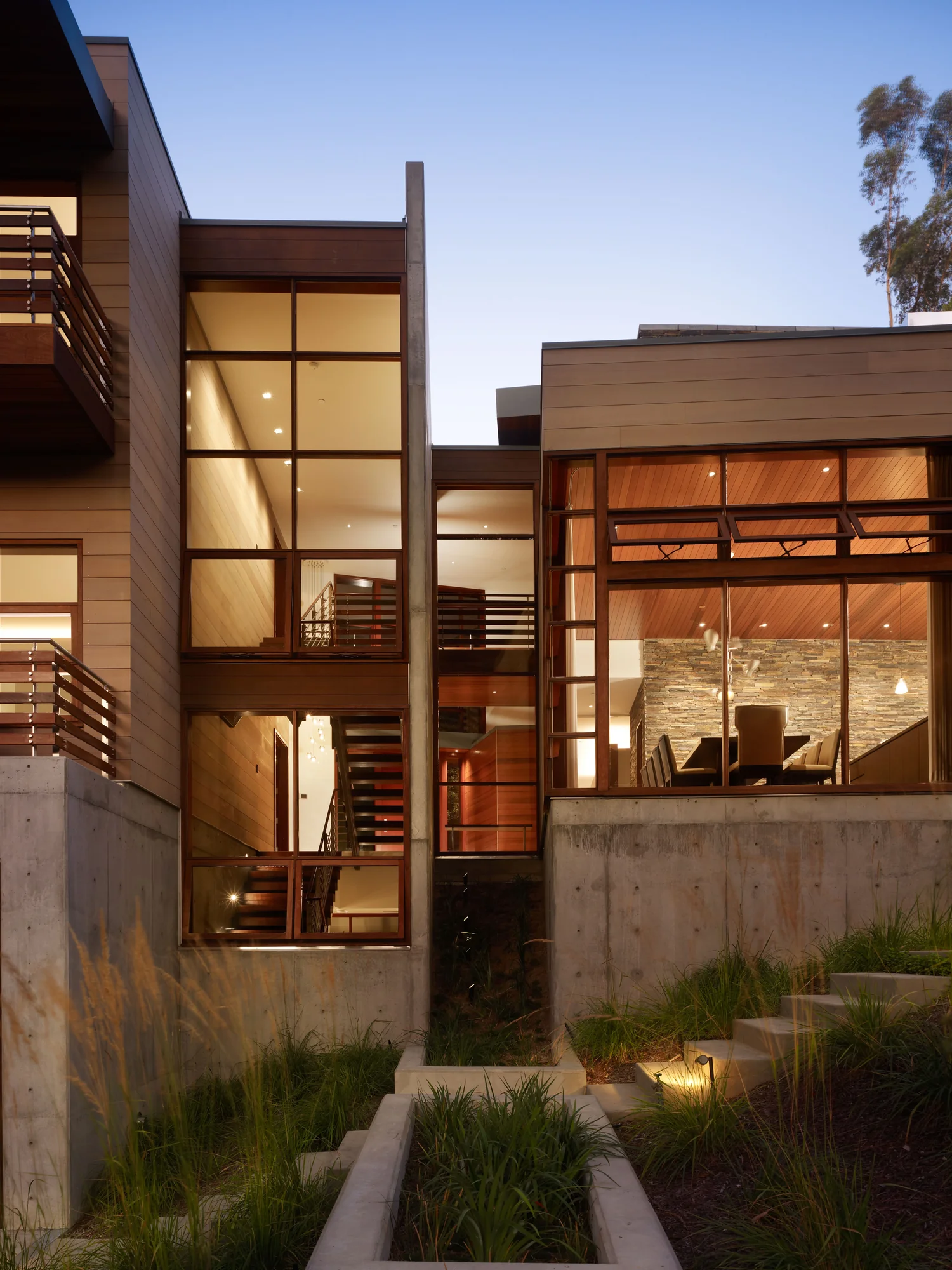 Exterior dusk view of a multilevel custom home with mahogany-framed windows, concrete base, and warmly lit interiors.