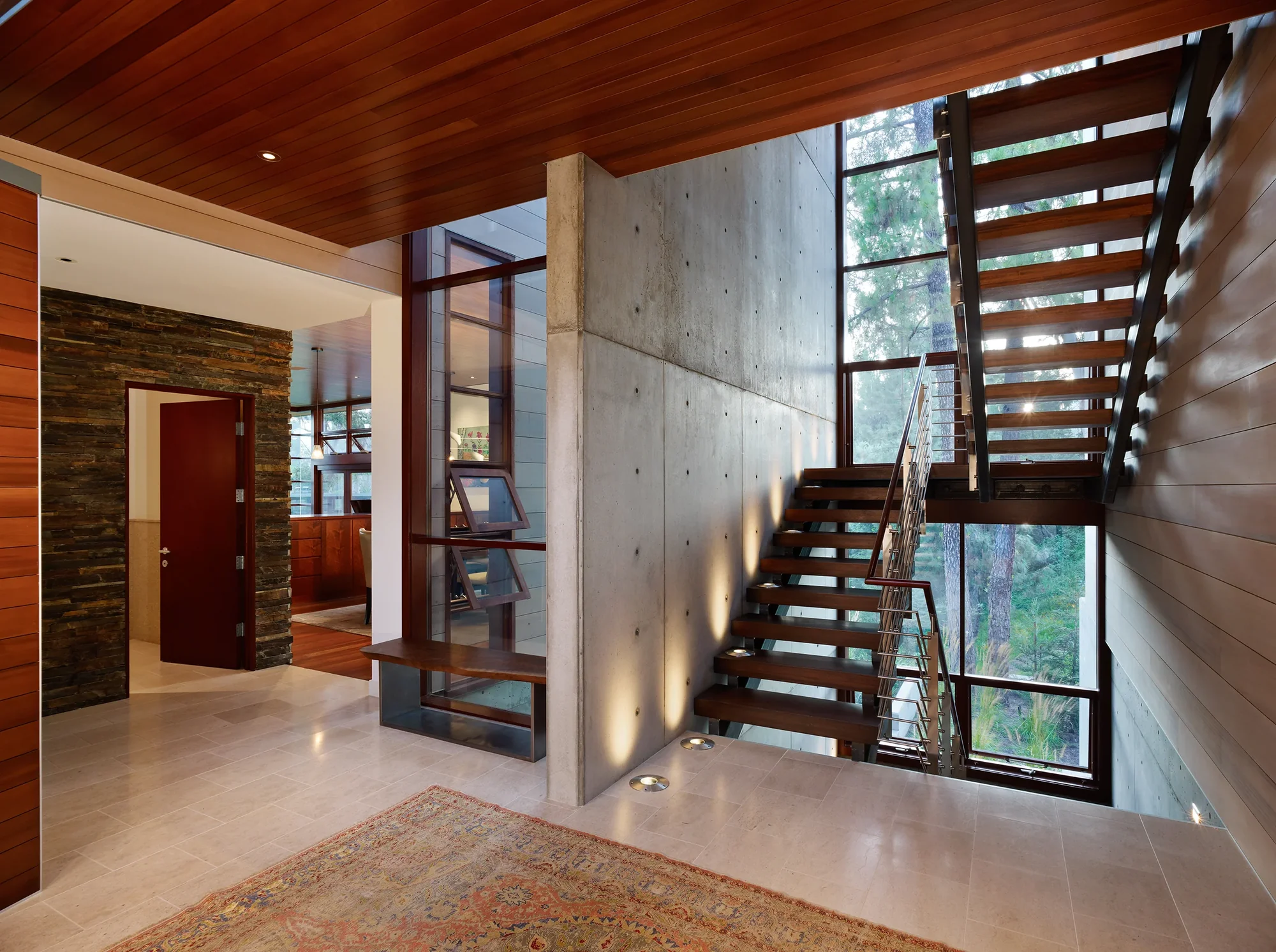 Floating mahogany staircase beside floor-to-ceiling windows framing forest views, with exposed concrete and warm wood ceiling.