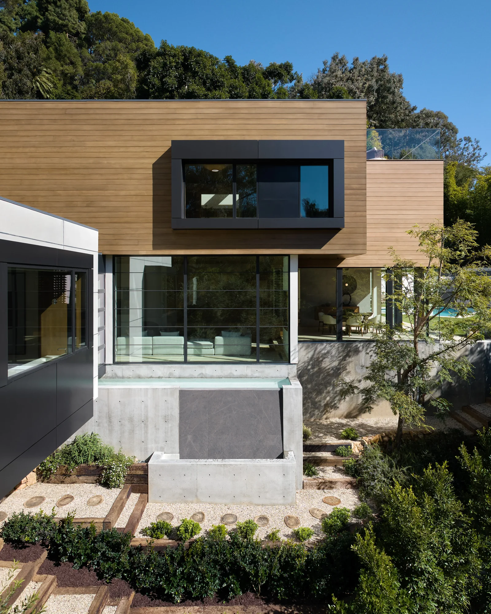 Modern home exterior featuring cedar siding, concrete base, and floor-to-ceiling glass walls opening to a landscaped garden.