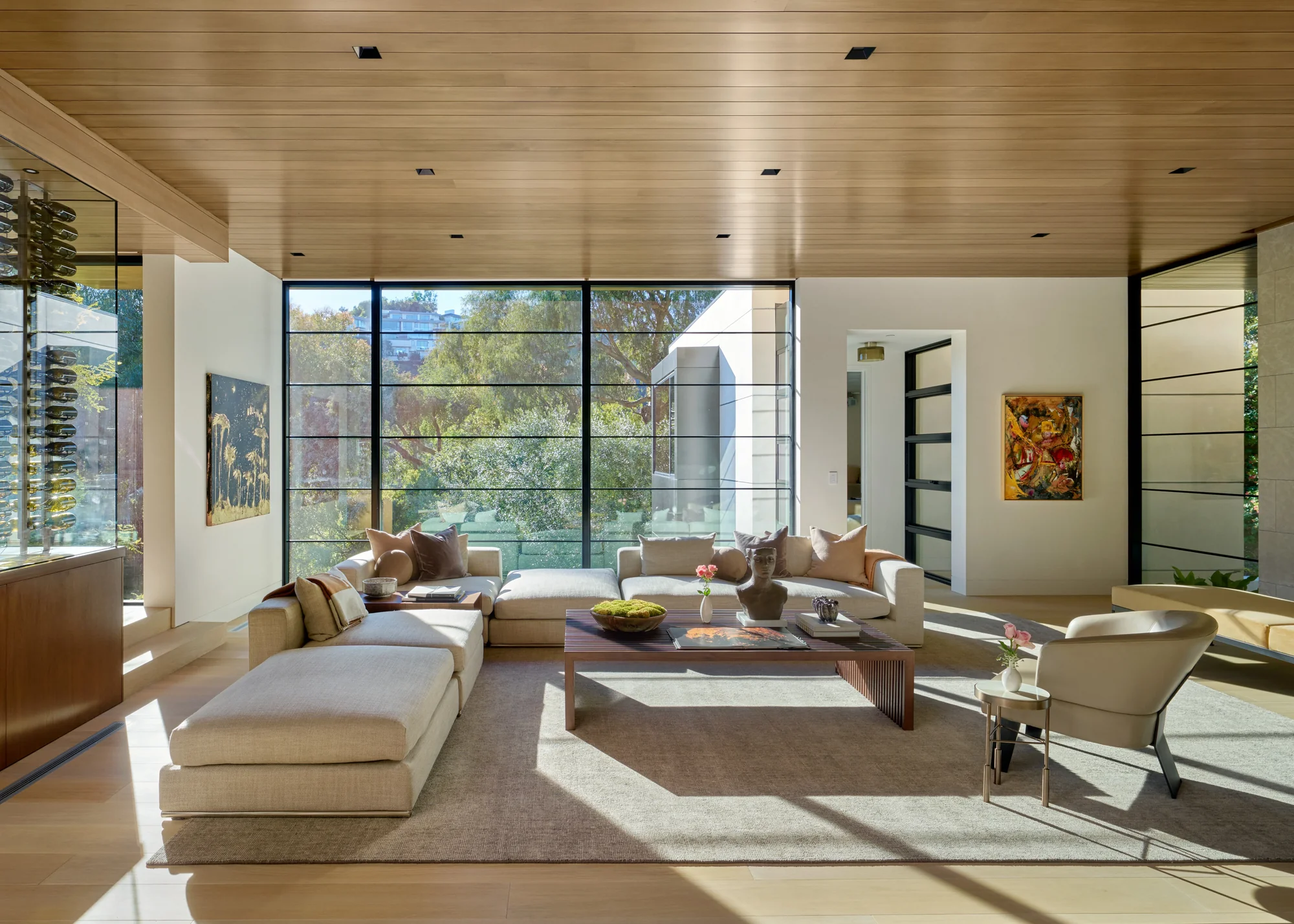 Sunlit living room with cedar ceiling, floor-to-ceiling steel windows, cream sectional, and lush tree views beyond.