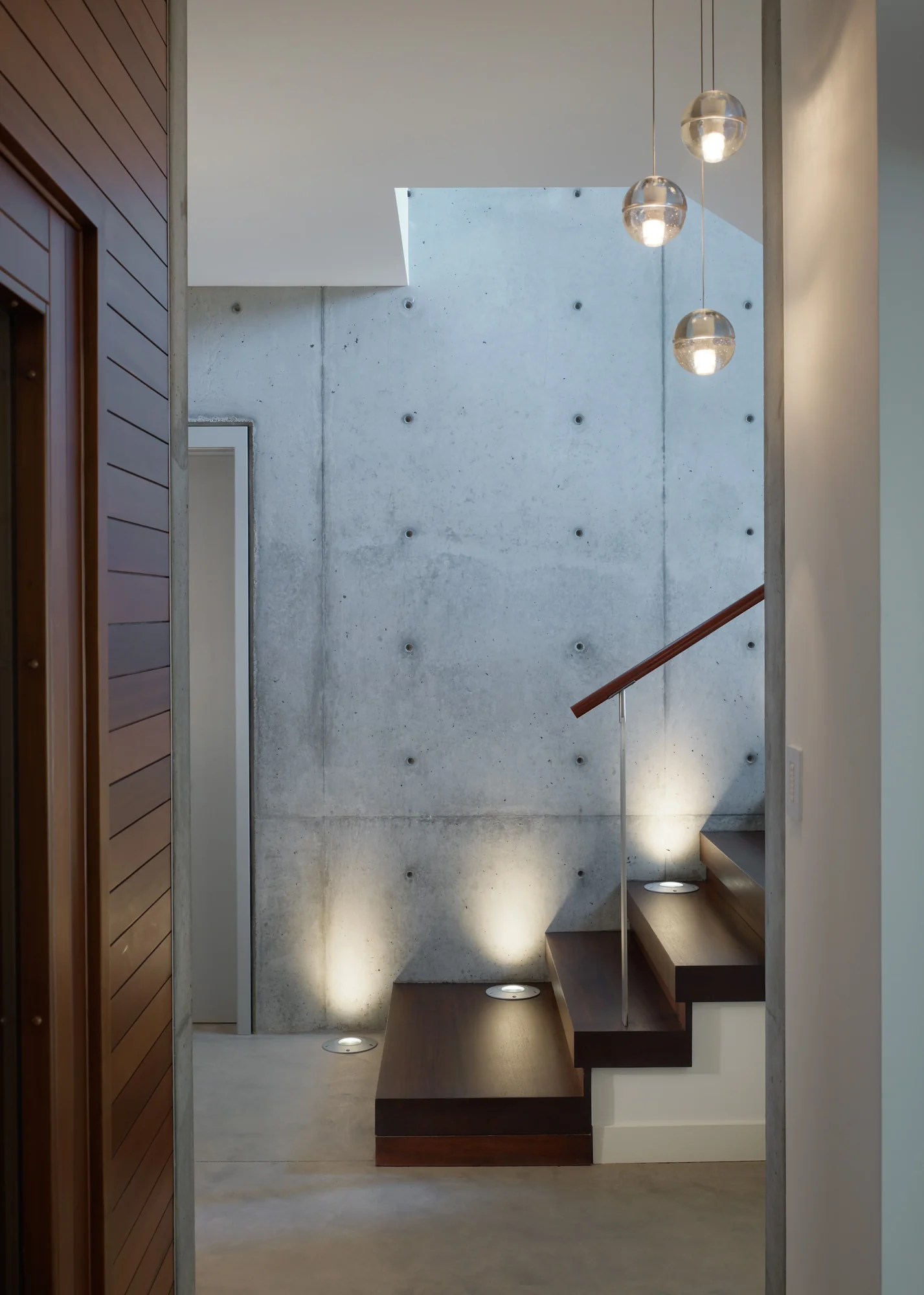 Floating mahogany staircase with exposed concrete wall, globe pendant lights, and recessed uplight fixtures in a modern entryway.