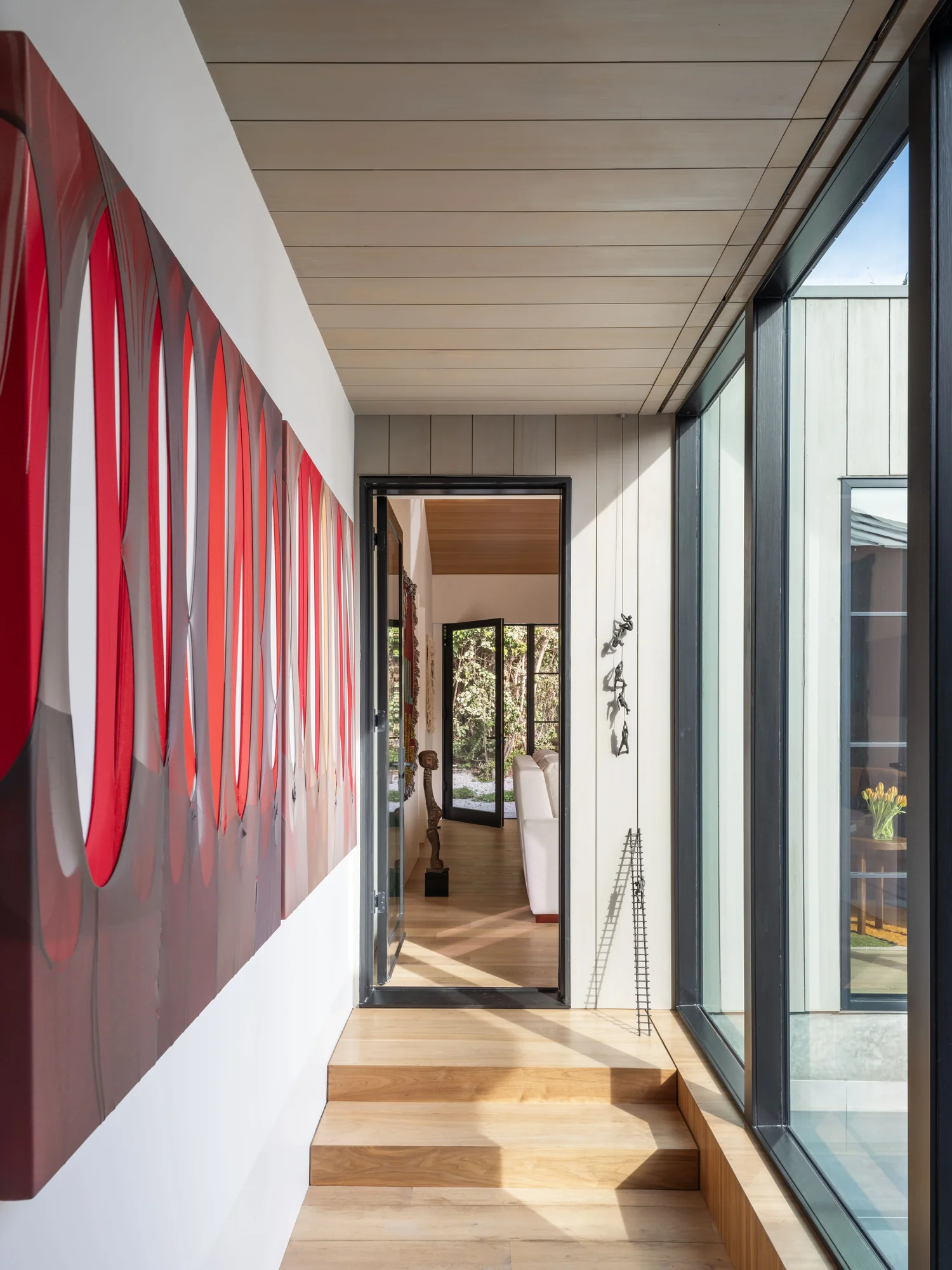 Modern hallway with light wood floors, floor-to-ceiling windows, red abstract art, and view through to exterior doors