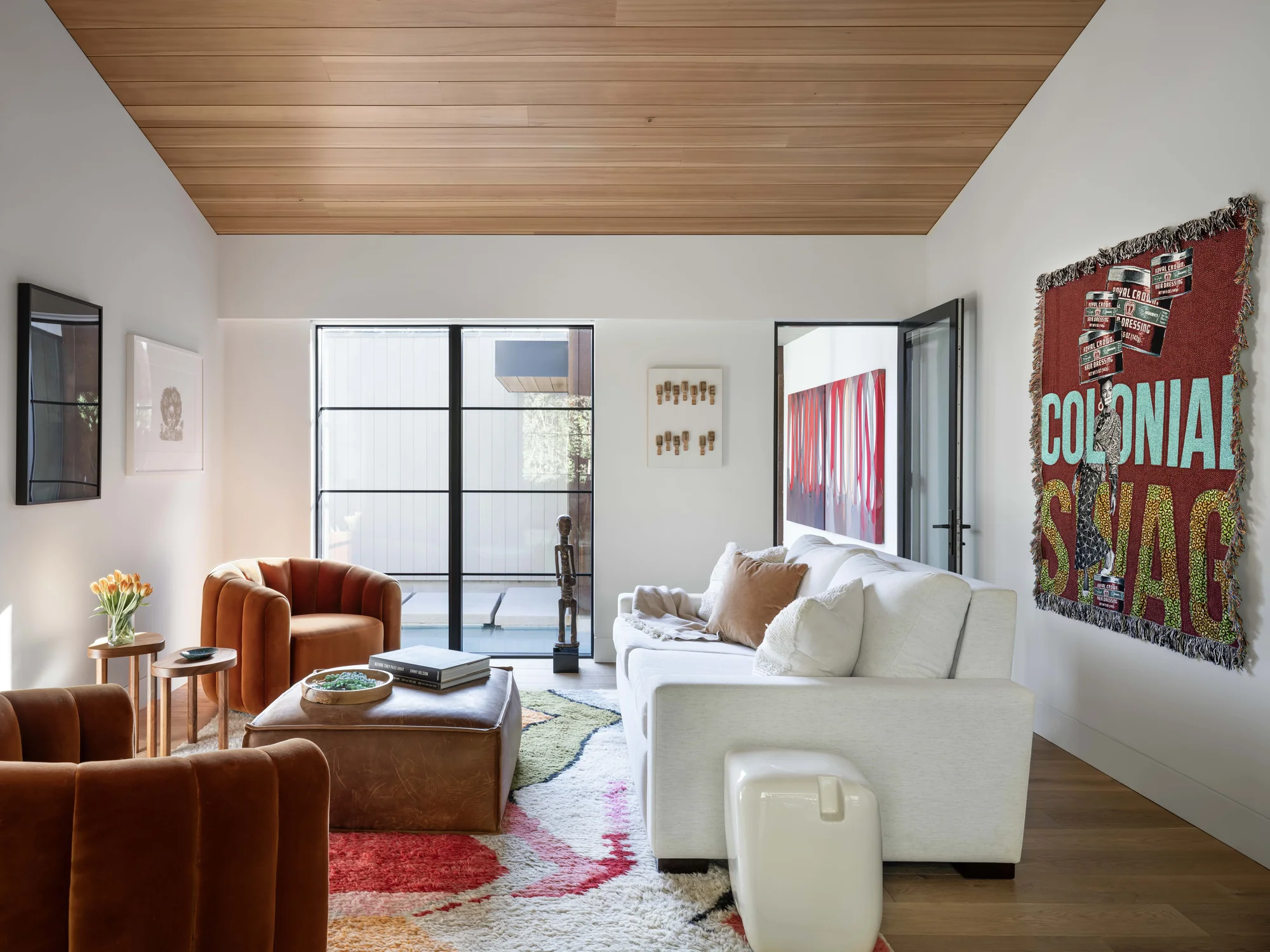 Modern living room with wood ceiling, white sofa, rust armchairs, glass doors opening to courtyard, and colorful art.