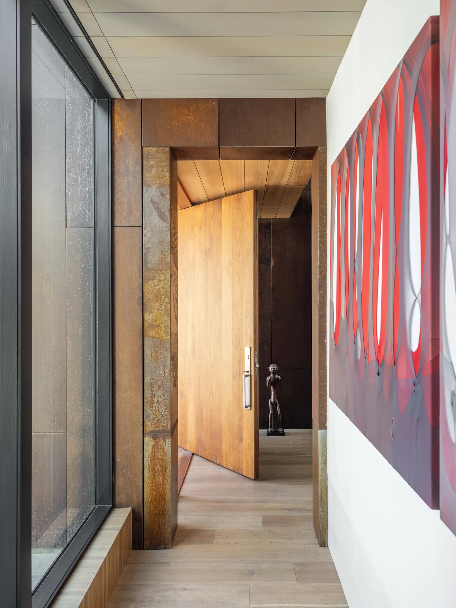 Modern entryway with pivoting wood door framed by weathered steel, light wood floors, and contemporary art on white wall.