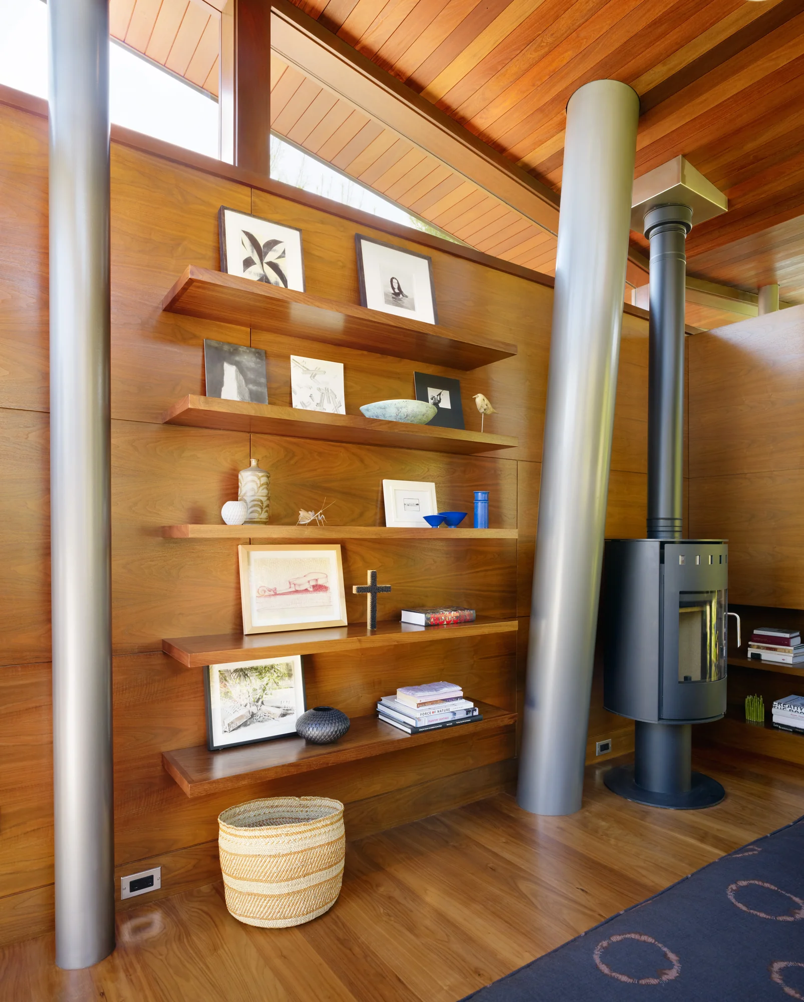 Warm wooden interior with floating shelves displaying art and books, modern wood stove, and cylindrical support columns