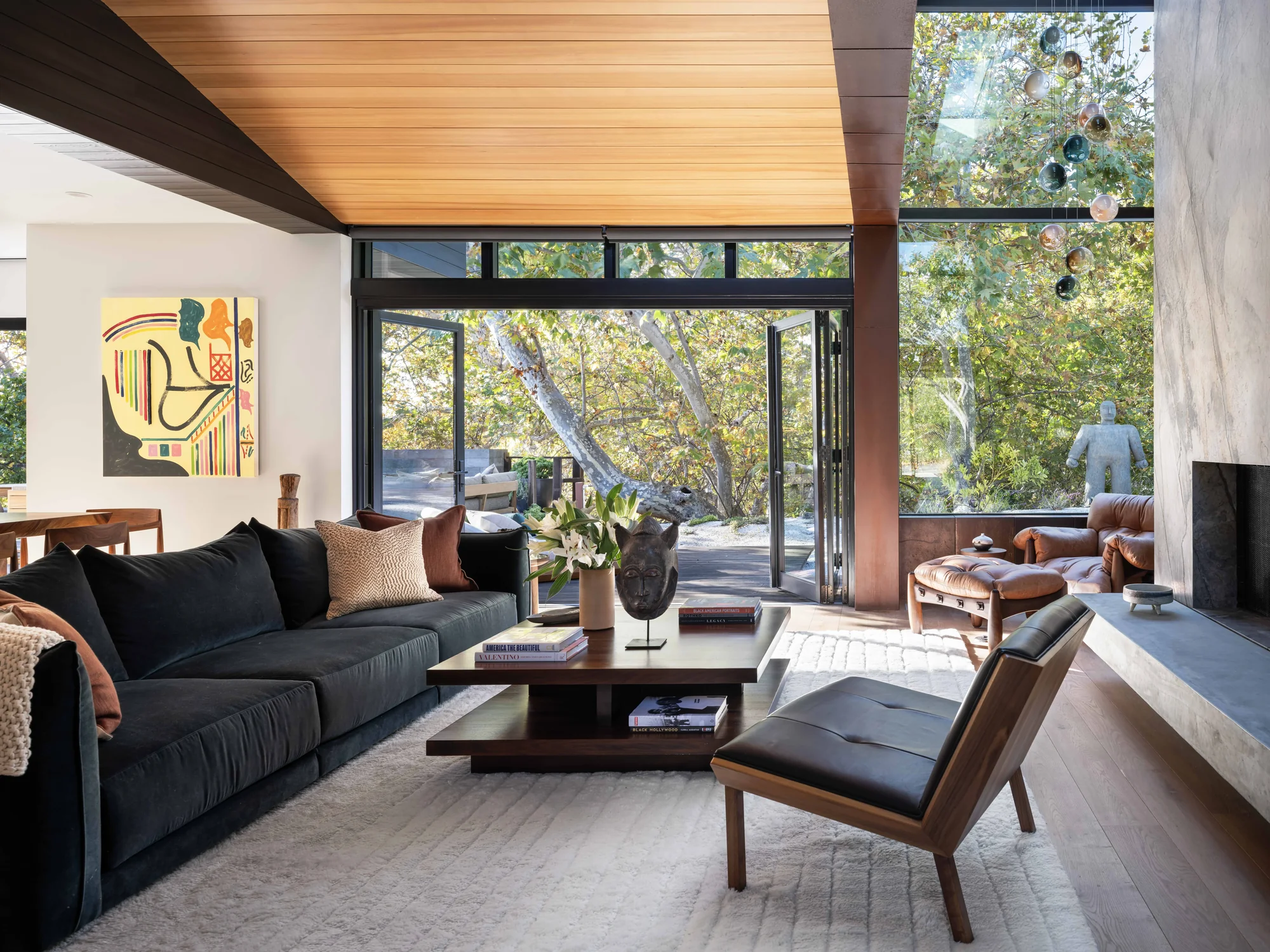 Modern living room with floor-to-ceiling windows opening to nature, featuring wood ceiling and contemporary furnishings.