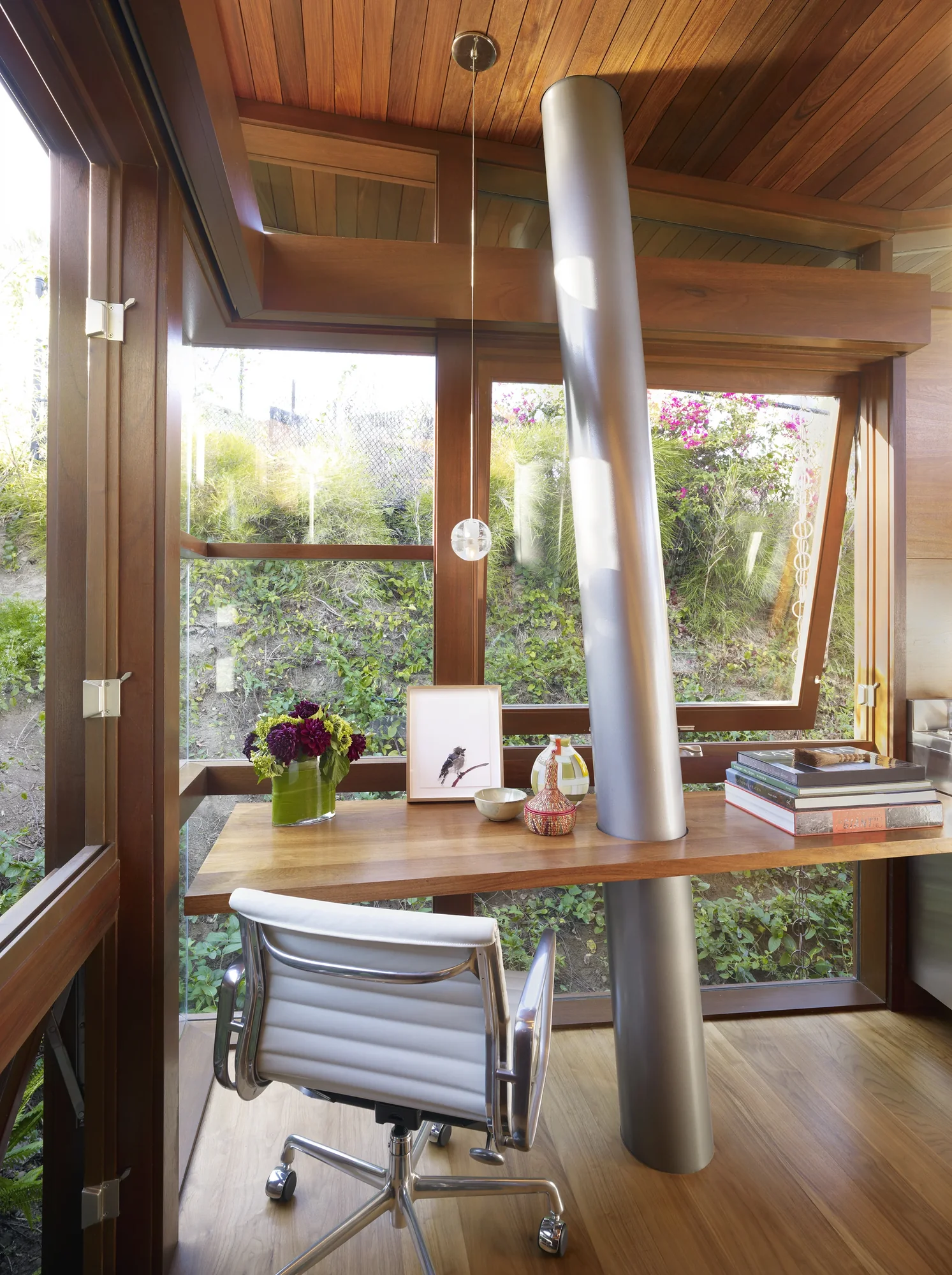 Modern wood-paneled home office with floor-to-ceiling windows overlooking garden, featuring desk, white leather chair, and cylindrical column.