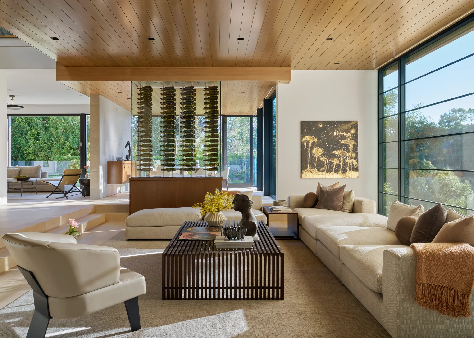Sunlit living room with cedar ceiling, wine display wall, cream sofas, and floor-to-ceiling windows overlooking lush greenery.
