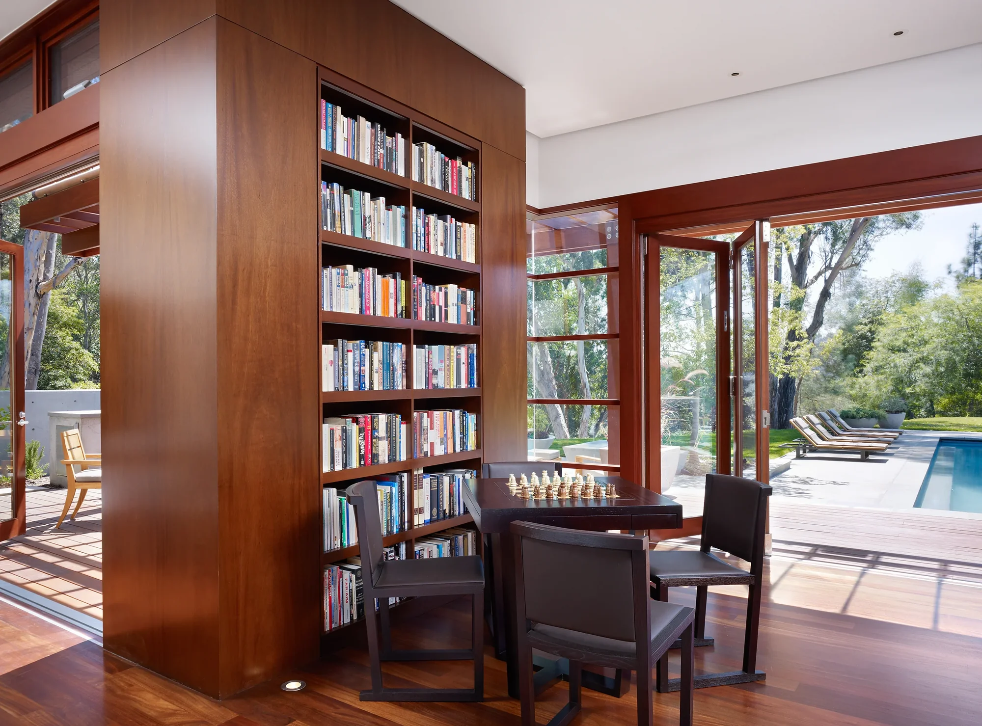 Built-in mahogany bookshelf with reading nook overlooking pool, featuring chess table and open views to outdoor living space